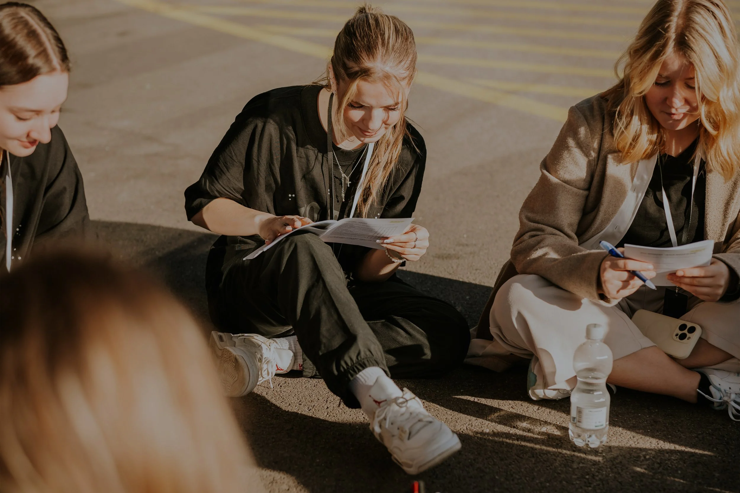 Drei junge Frauen sitzen auf dem Boden, lesen Papierdokumente und haben Wasserflaschen neben sich.