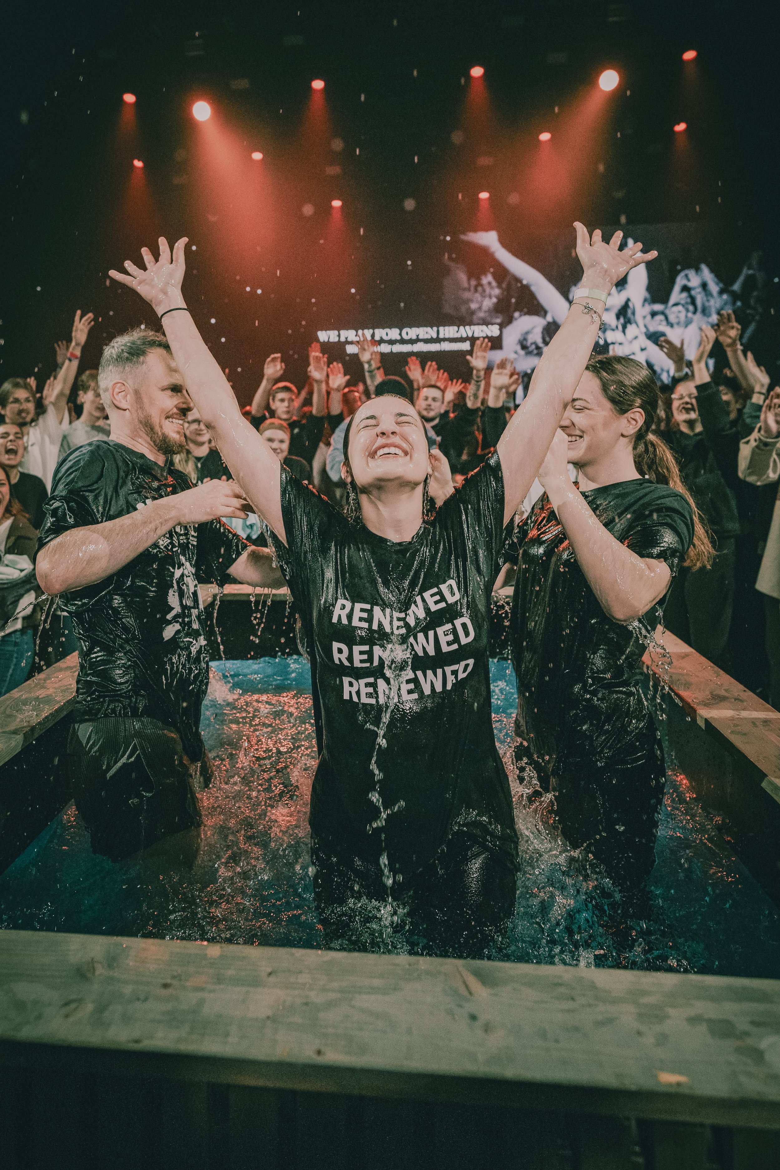 Eine jubelnde Frau mit erhobenen Händen steht im Wasser, umgeben von anderen Menschen in einem Veranstaltungsraum. Im Hintergrund sind Lichter und eine große Leinwand mit Text zu sehen.