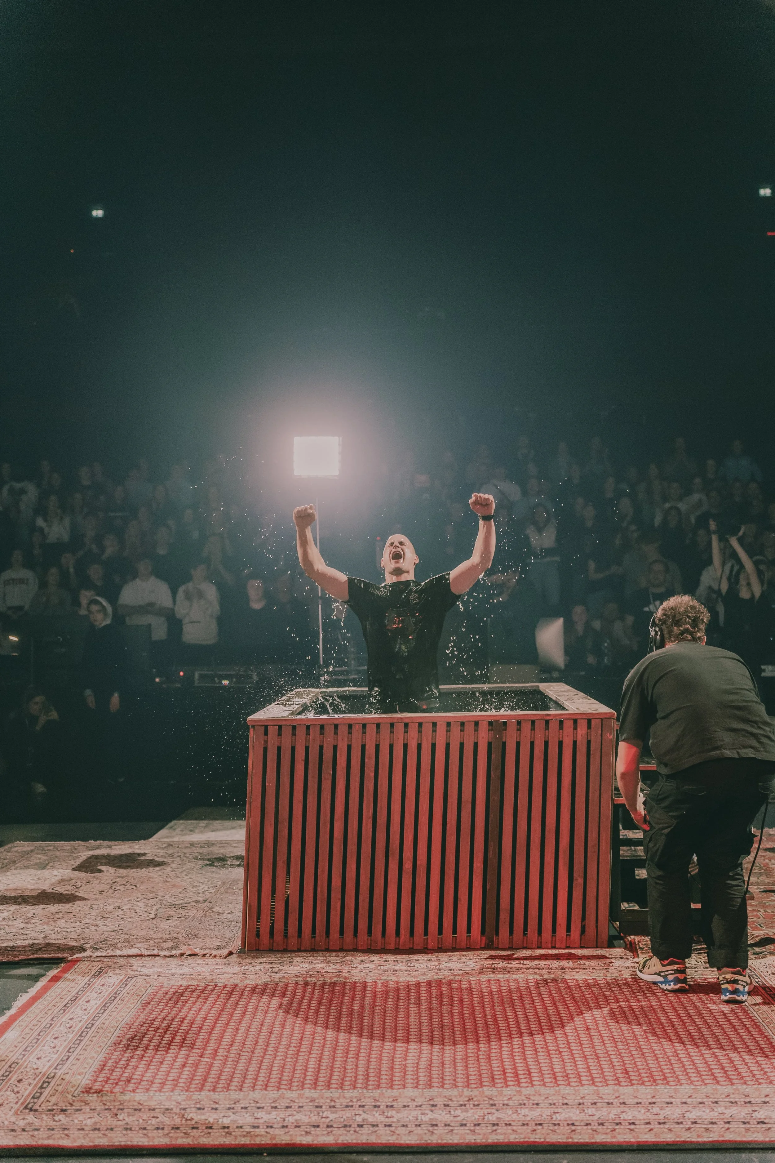 Ein Mann in einem dunklen T-Shirt jubelt in einer großen Menschenmenge, während er in einem Wasserbecken steht, aus dem Wasser spritzt.