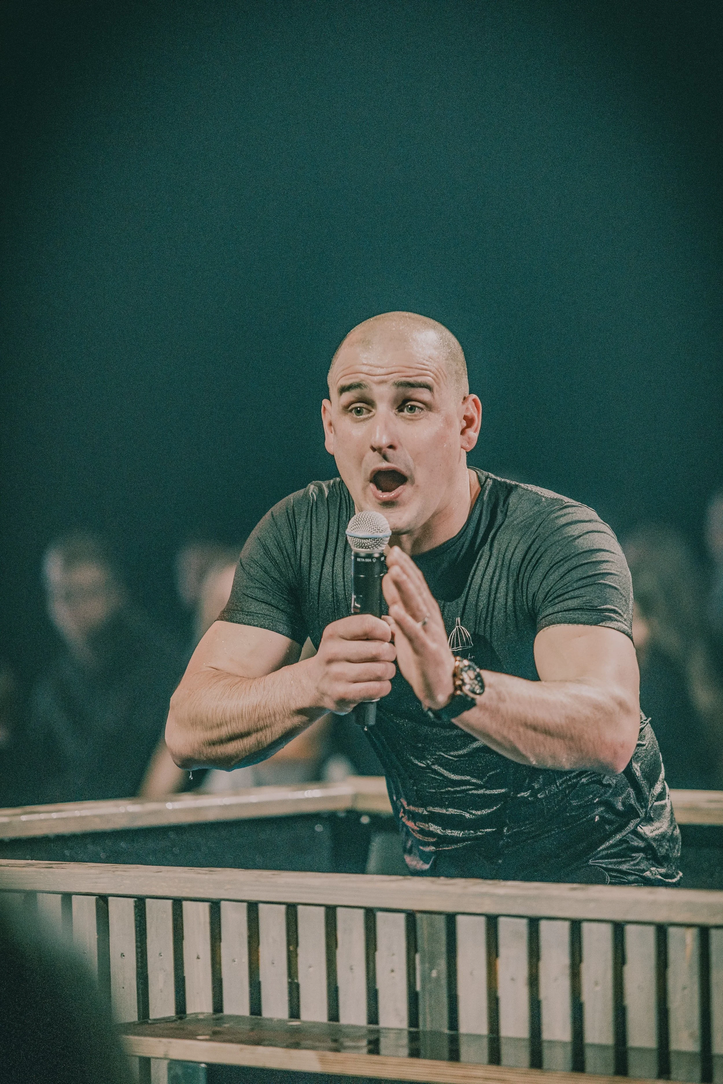 Mann im schwarzen T-Shirt spricht in ein Mikrofon, während er sich auf einer Bühne befindet.