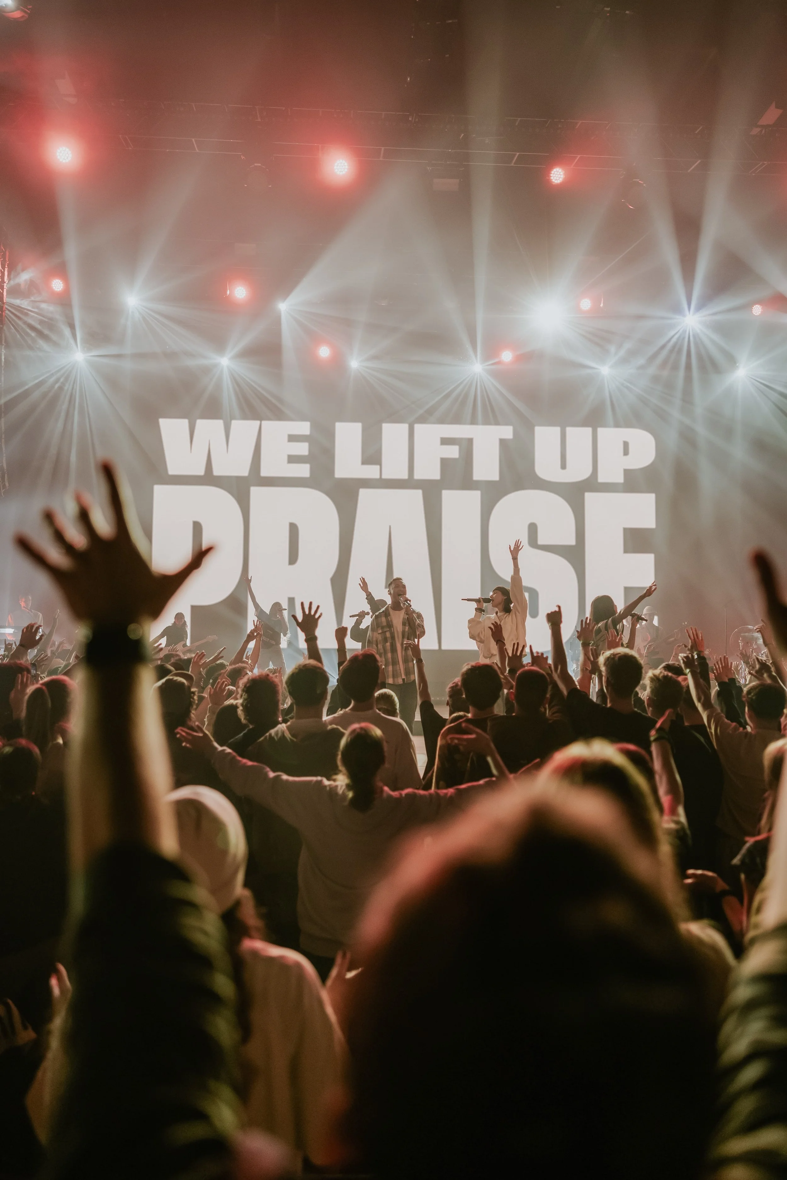 Eine Menschenmenge bei einem Konzert mit aufgerissenen Händen, im Vordergrund sind Personen mit erhobenen Armen, im Hintergrund auf der Bühne sind Sänger mit Mikrofonen, vor einer großen leinwand, auf der in großen Buchstaben 'WE LIFT UP PRAISE' steht, buntes Bühnenlicht und Scheinwerfer.