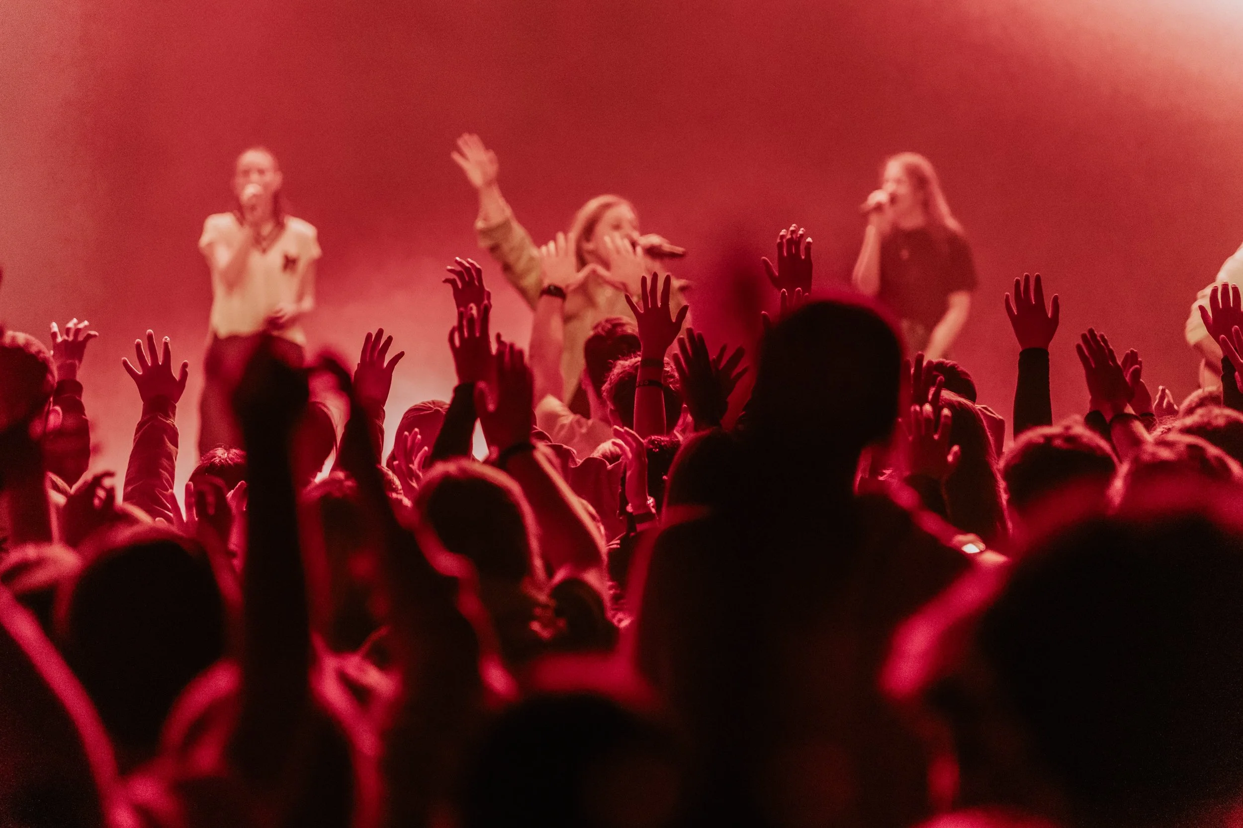 Menschenmenge auf Konzert, Hände in die Luft, Bühne mit Sängerinnen im Hintergrund, rotes Licht.