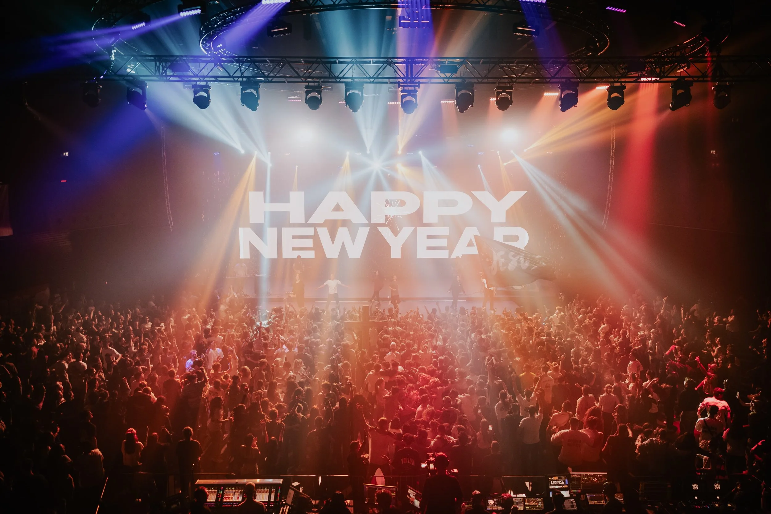 Eine große Menschenmenge tanzt auf einer Feier im Innenraum. Auf einer großen Leinwand im Hintergrund steht in weißen Buchstaben 'HAPPY NEW YEAR'. Es gibt bunte Lichtstrahlen, die auf die Menge gerichtet sind, und die Atmosphäre ist festlich.