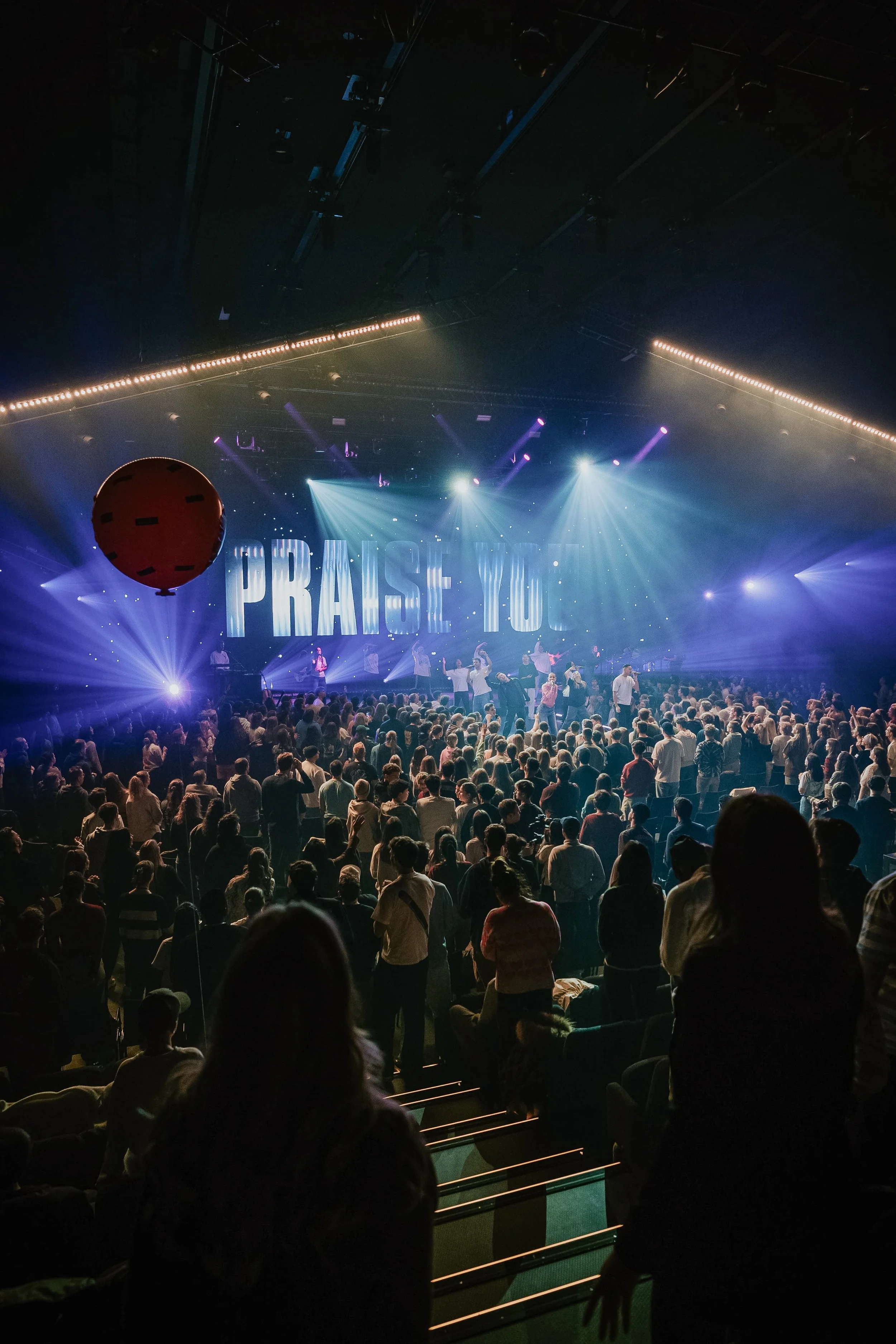 Menschenmenge in einer Kirche oder bei einem religiösen Event, das auf der Bühne die Worte "PRAISE YOU" zeigt, mit bunten Lichtern und einer großen roten Ballons.