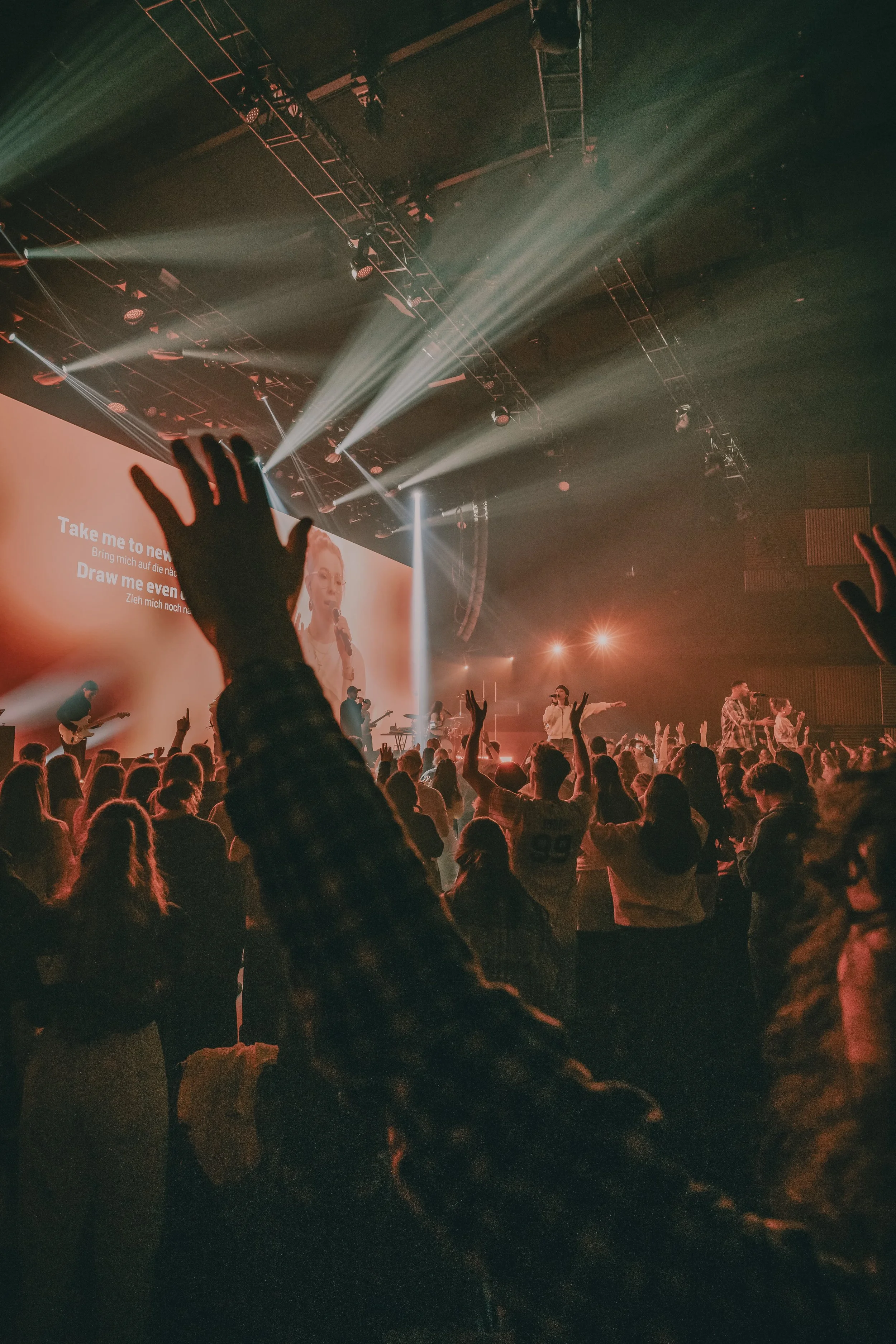 Menschen in einem dunklen Konzertsaal mit Scheinwerfern und einer großen Leinwand, auf der Karaoke-Texte in Englisch und Deutsch angezeigt werden.