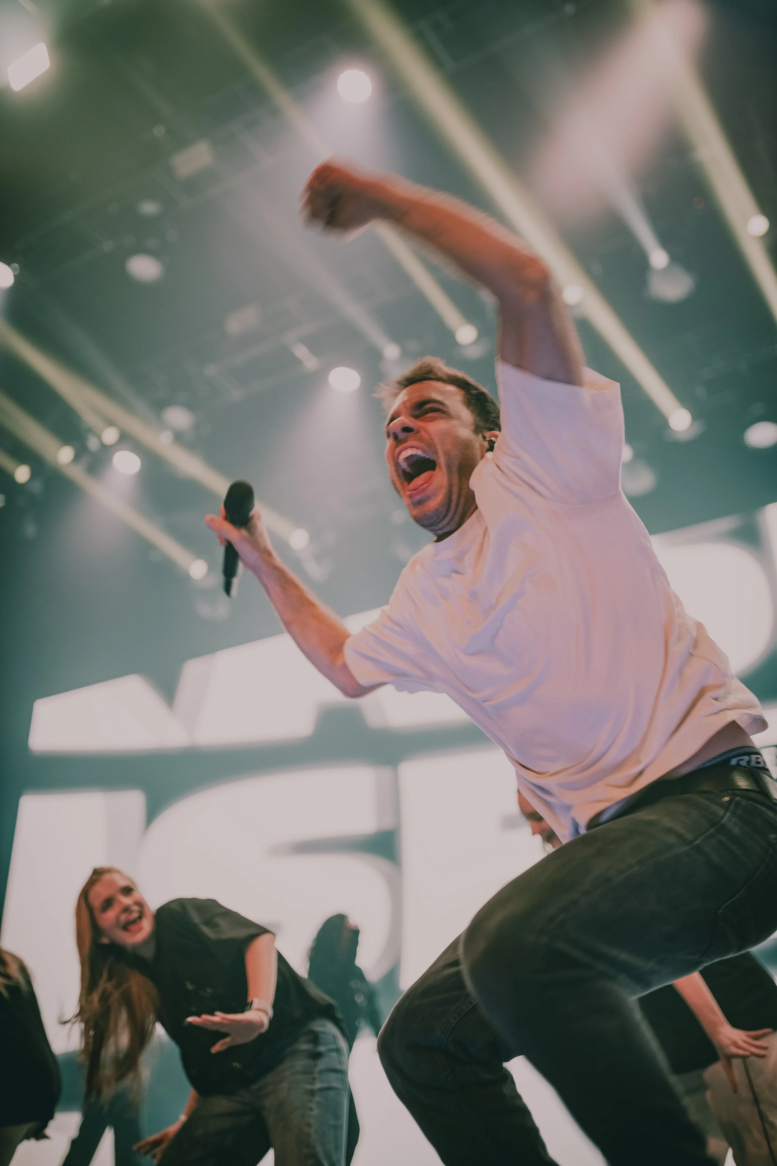 Junger Mann singt und tanzt auf einer Bühne mit Mikrofon, umgeben von mehreren lachenden Menschen, bei einer Veranstaltung oder Show.