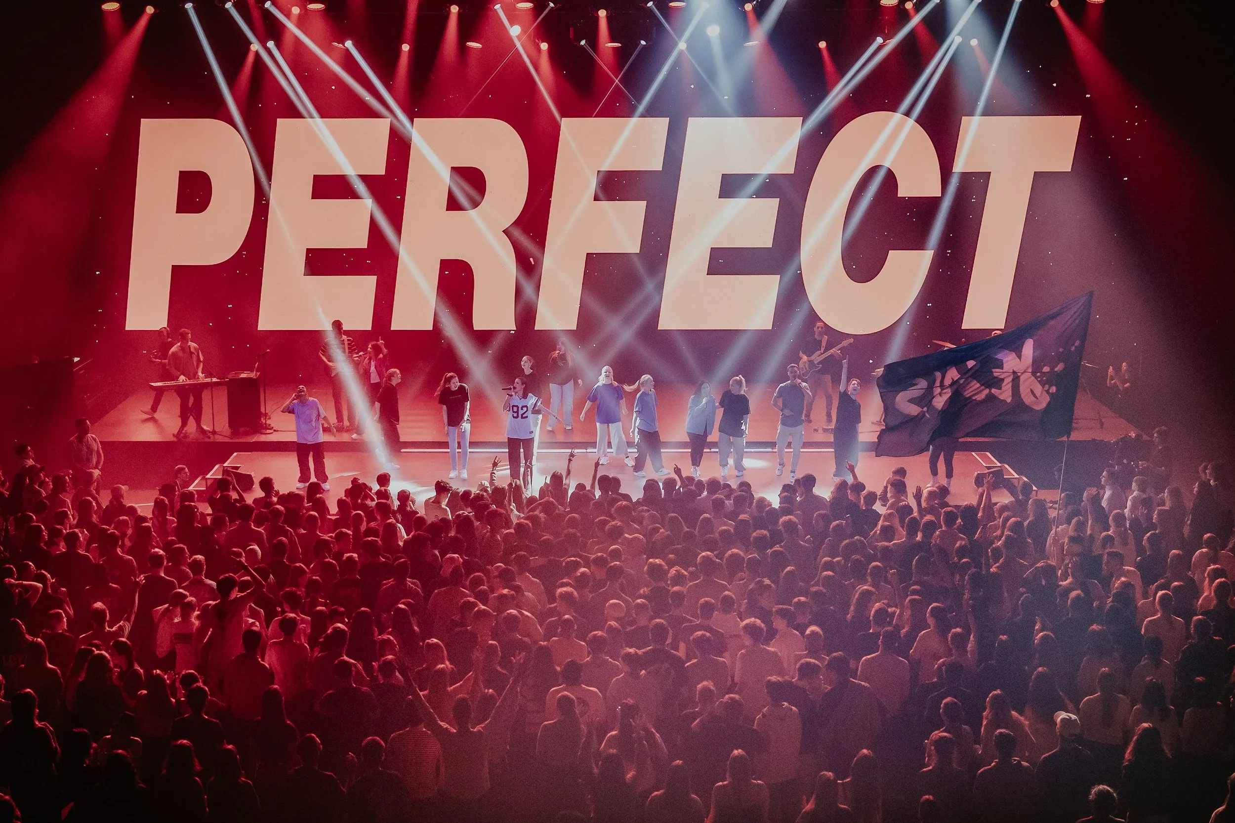 Auf einer Bühne im Konzertsaal stehen mehrere Sänger und Musiker, während im Hintergrund das Wort 'PERFECT' groß projiziert wird. Das Publikum ist in Erwartung, einige halten Flaggen hoch, es gibt bunte Lichter und eine lebhafte Atmosphäre.
