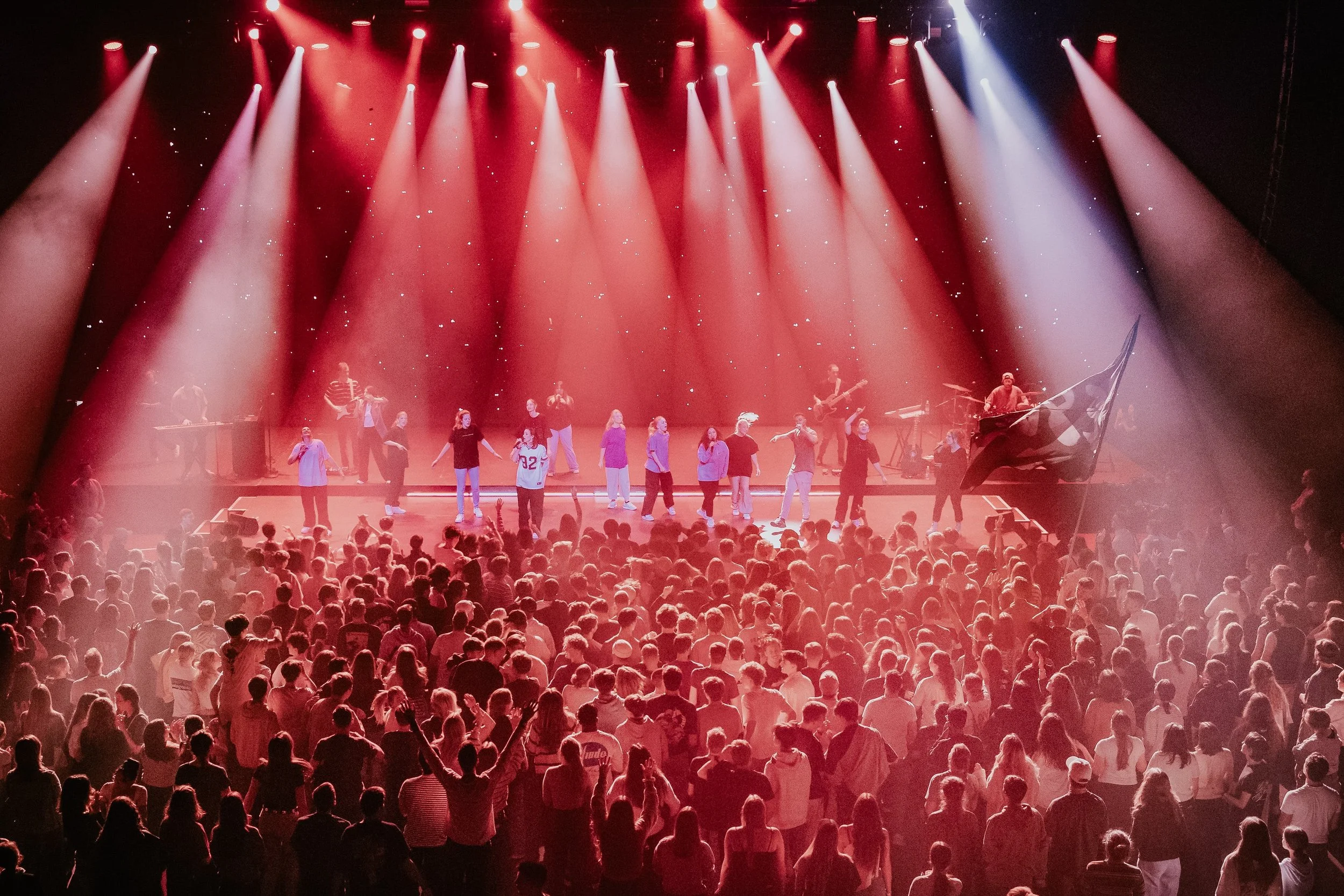 Eine Bühne mit mehreren Sängern und Musikern, die von roten Scheinwerfern beleuchtet wird, vor einem Publikum bei einer Konzert- oder Showveranstaltung.