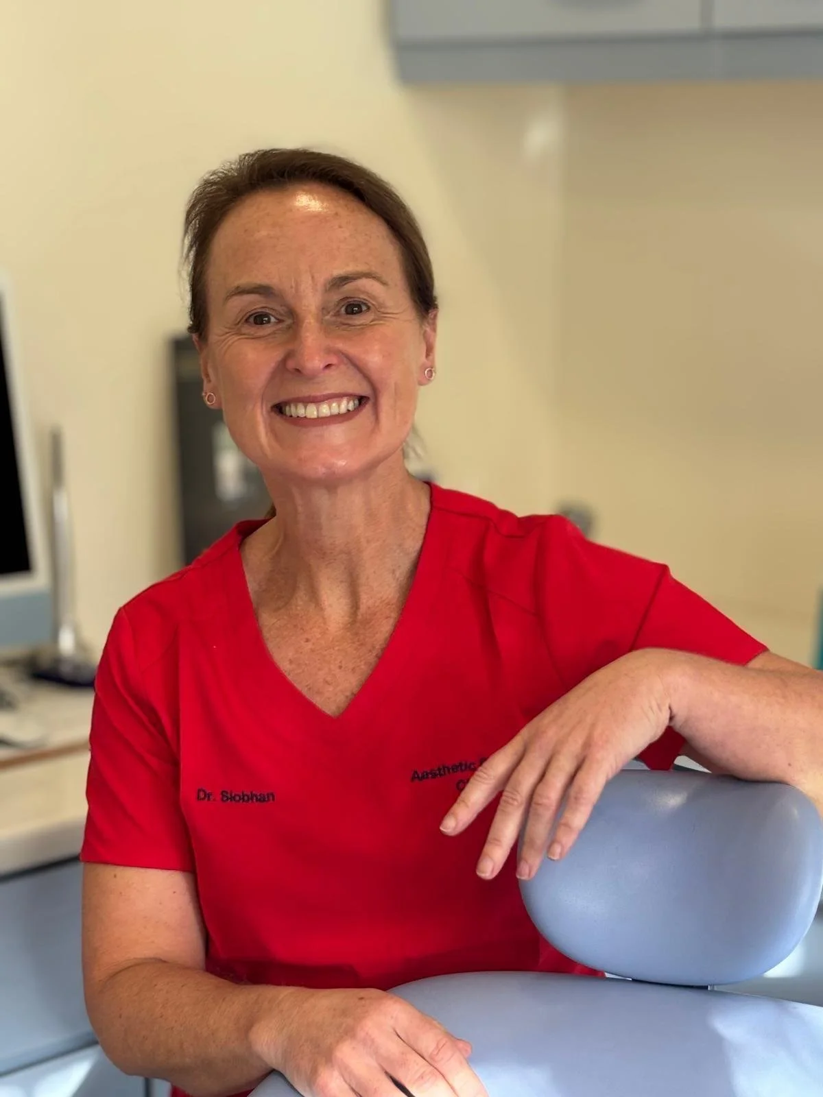 A woman with short brown hair, smiling and wearing a red medical uniform, seated in a clinic or hospital setting.