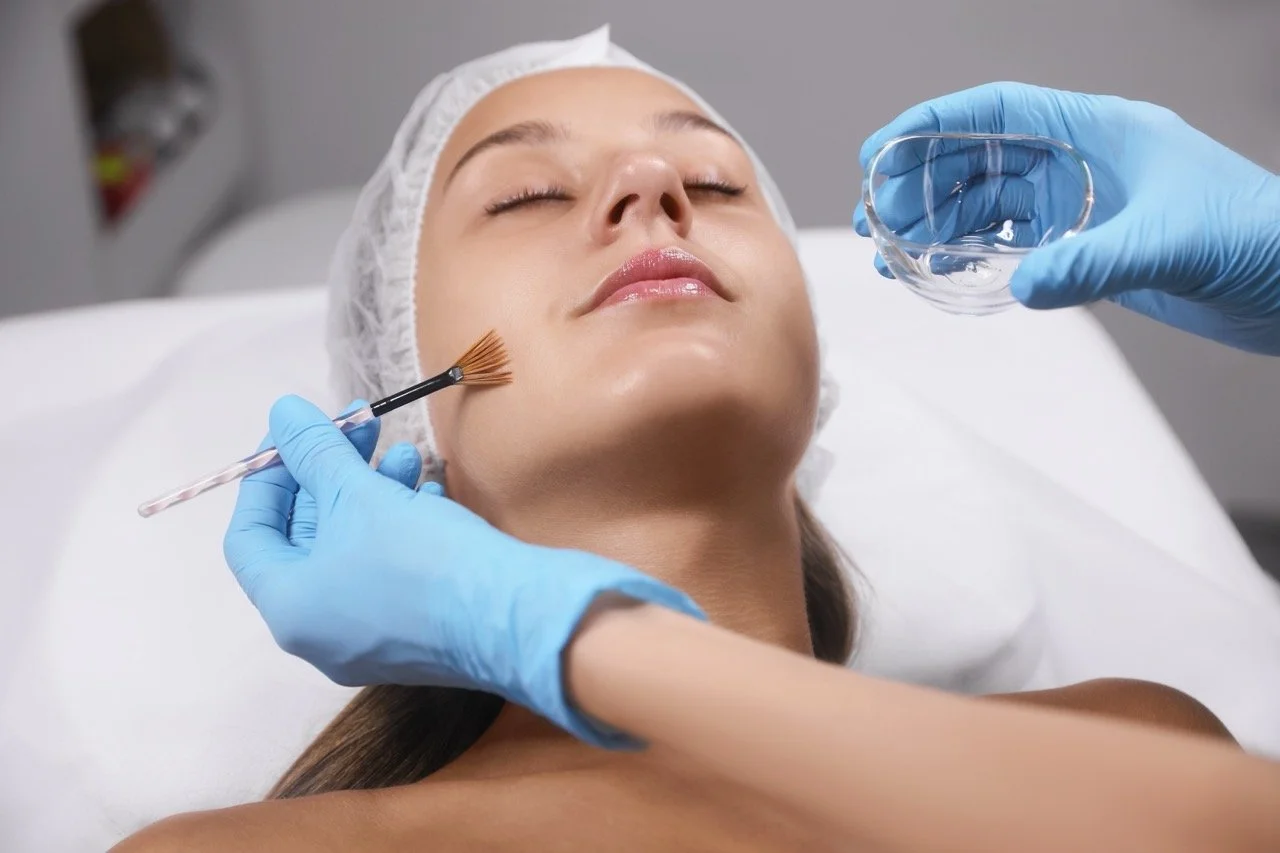 A woman receiving a facial treatment in a medical or spa setting, with a healthcare professional wearing gloves applying a skincare product with a brush while holding a small glass bowl.