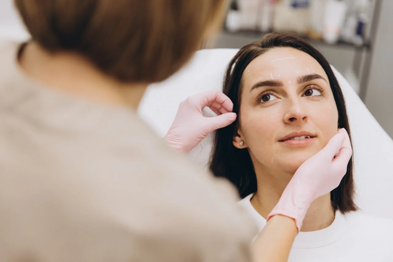 A woman receiving a cosmetic consultation or facial treatment from a healthcare professional in a clinical setting.