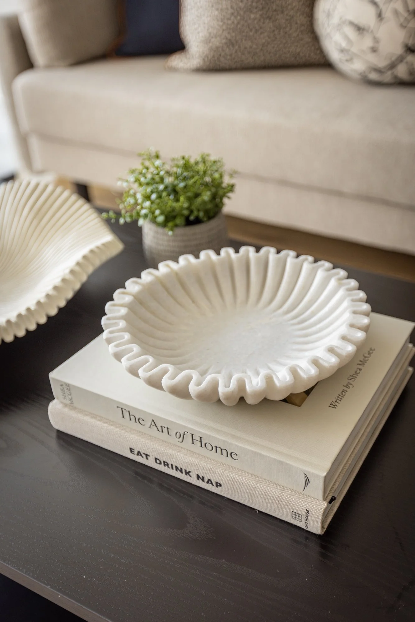 Decorative white ceramic bowl with a ruffled edge placed on top of two stacked books, one titled 'The Art of Home' and the other 'Eat Drink Nap', on a dark wooden surface, with a beige couch and potted green plant in the background.