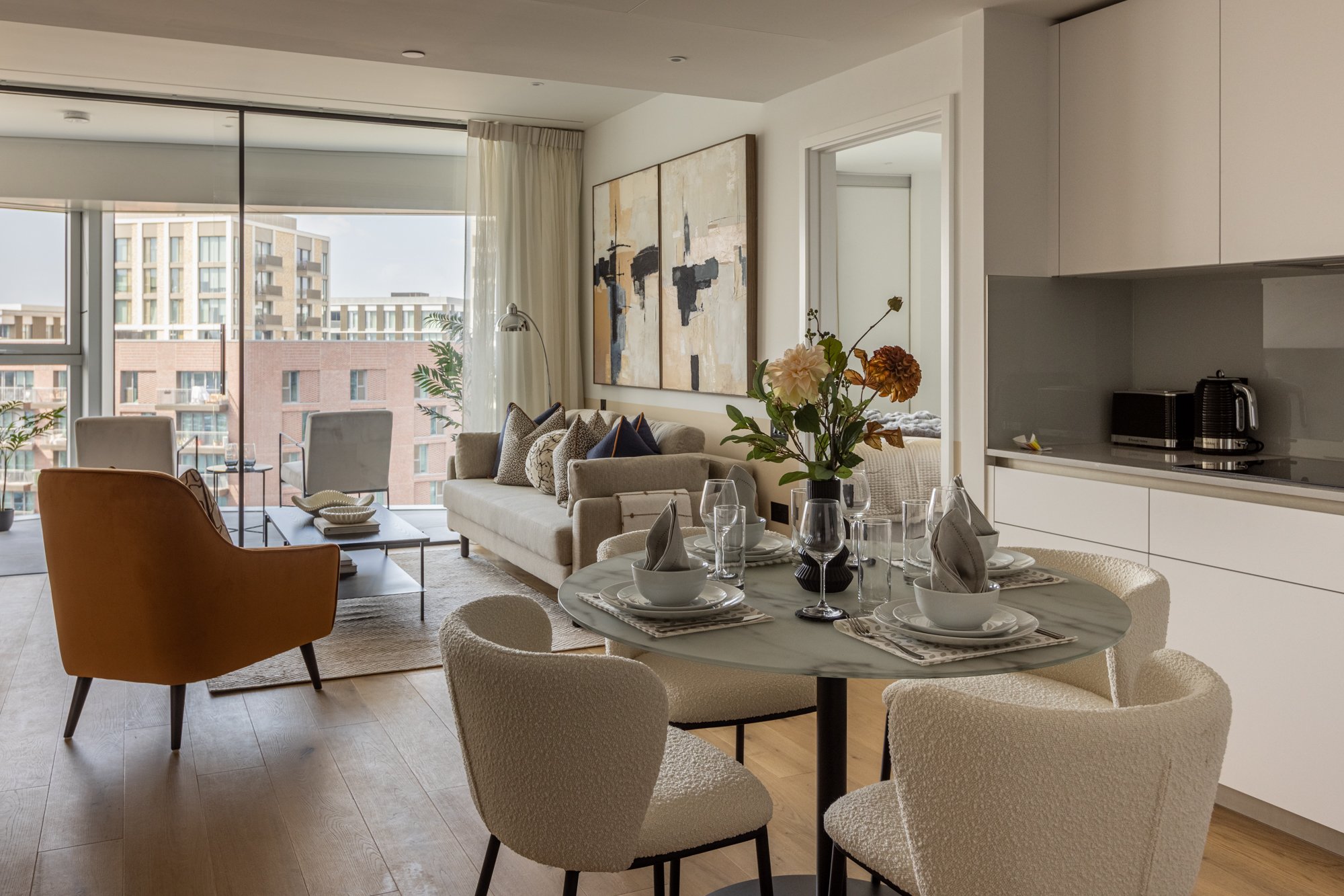 Modern living and dining area with a round marble table set for four, a vase with flowers, a beige couch with decorative pillows, an orange armchair, large windows with balcony views, and a kitchen area with white cabinets and small appliances.
