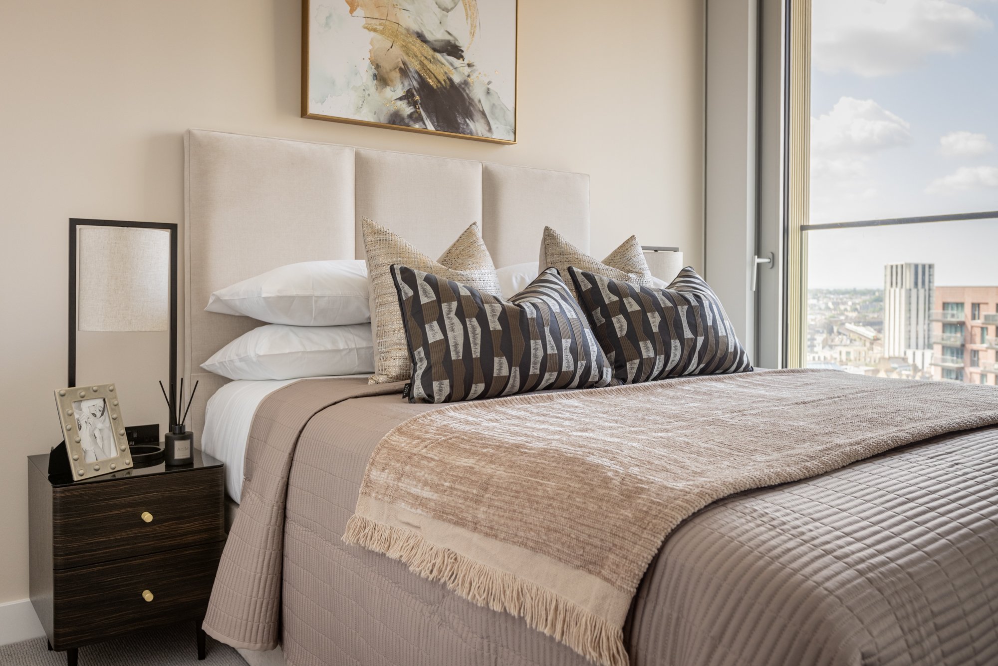 Modern bedroom with a large window, white upholstered headboard, abstract art above the bed, and decorative pillows.