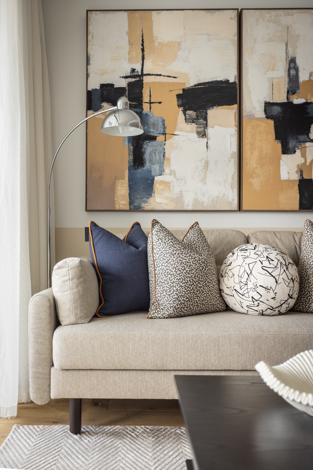 Living room with a beige sofa decorated with three pillows: a solid navy pillow, a small patterned pillow, and a large white pillow with black abstract lines. Behind the sofa, two large abstract paintings with black, white, beige, and blue tones hang