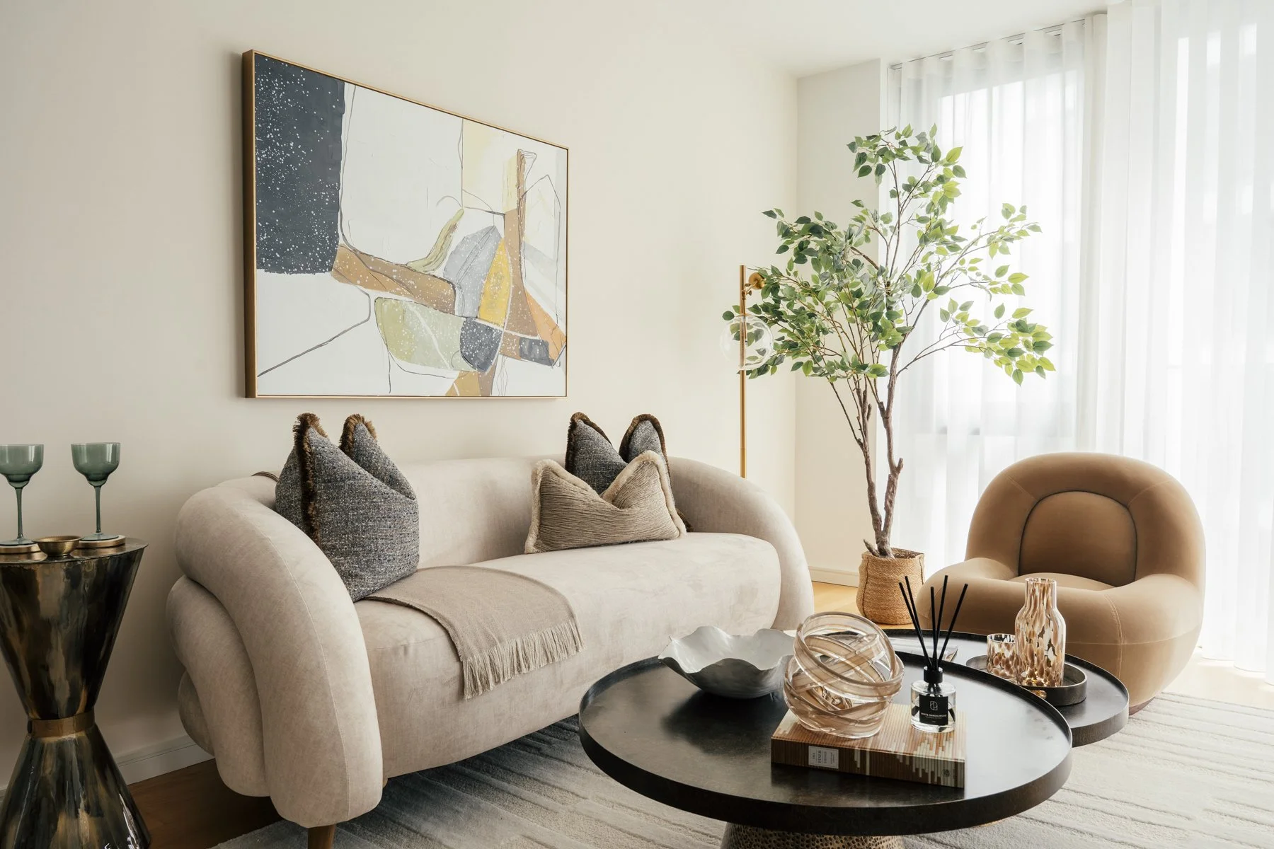 A modern living room with a beige sofa, decorative pillows, a round coffee table with decorative items, a tan armchair, a large potted plant, and abstract wall art, with sheer curtains allowing natural light.
