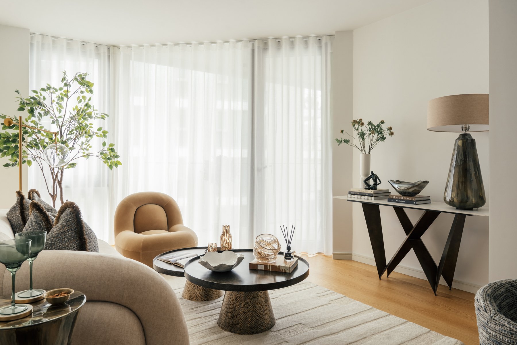 Bright living room with beige and brown furniture, a round black coffee table with decorative items, sheer white curtains, and a black console table with accessories and a large table lamp.