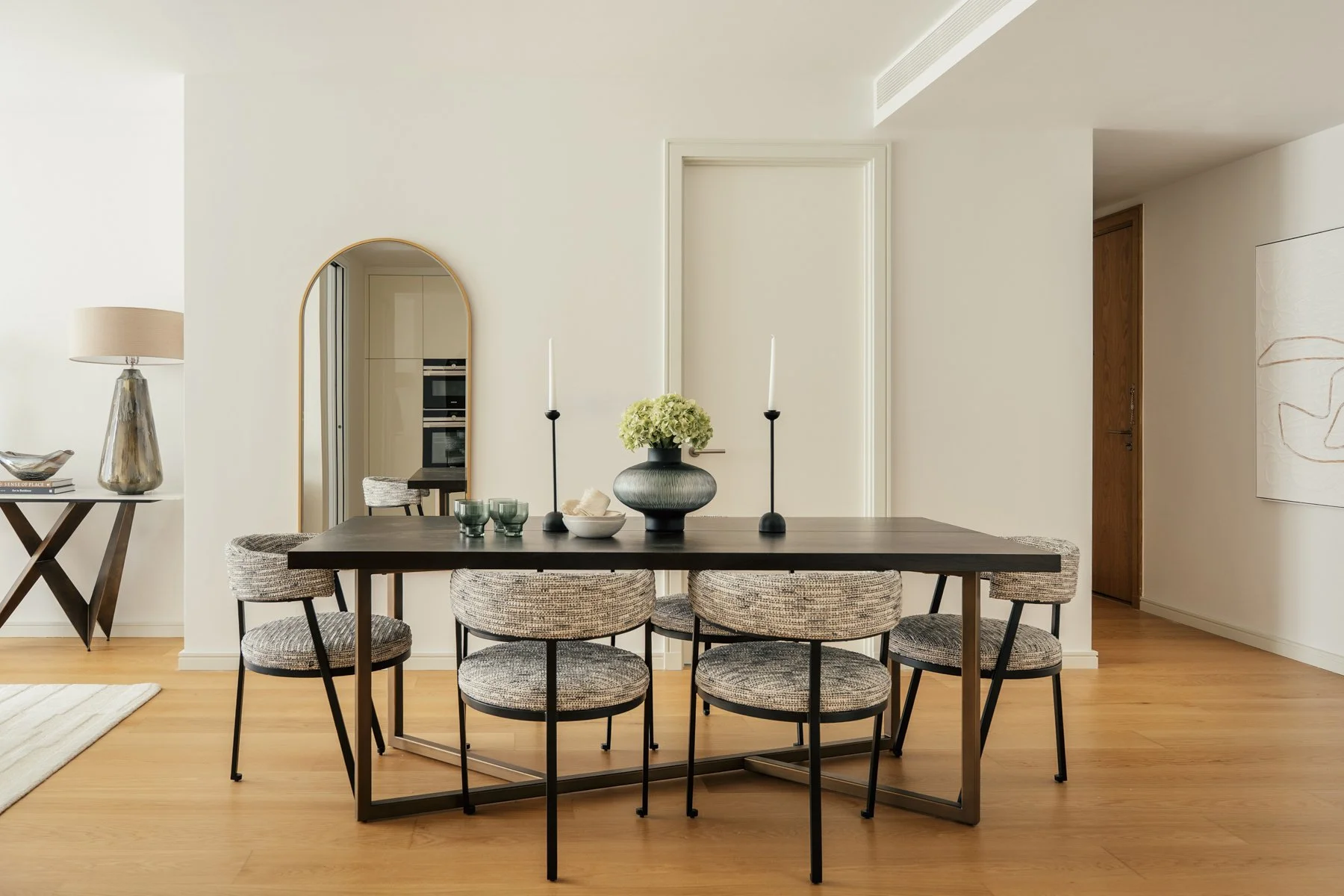 Black dining table with six upholstered chairs, decorative vase with green flowers, and two tall candle holders on a modern wooden floor in a bright, minimalist dining room.