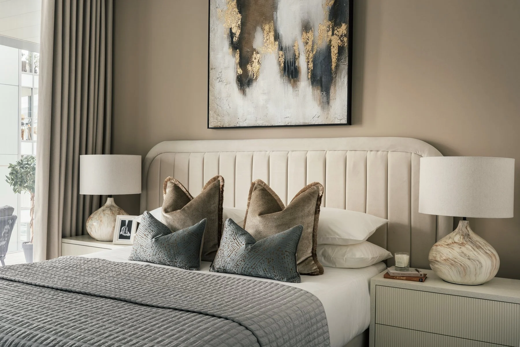 A bedroom with a white upholstered headboard, beige walls, and a large abstract painting above the bed. The bed has multiple pillows in shades of brown and teal, and a gray quilted bedspread. On each side of the bed, there are white nightstands with 