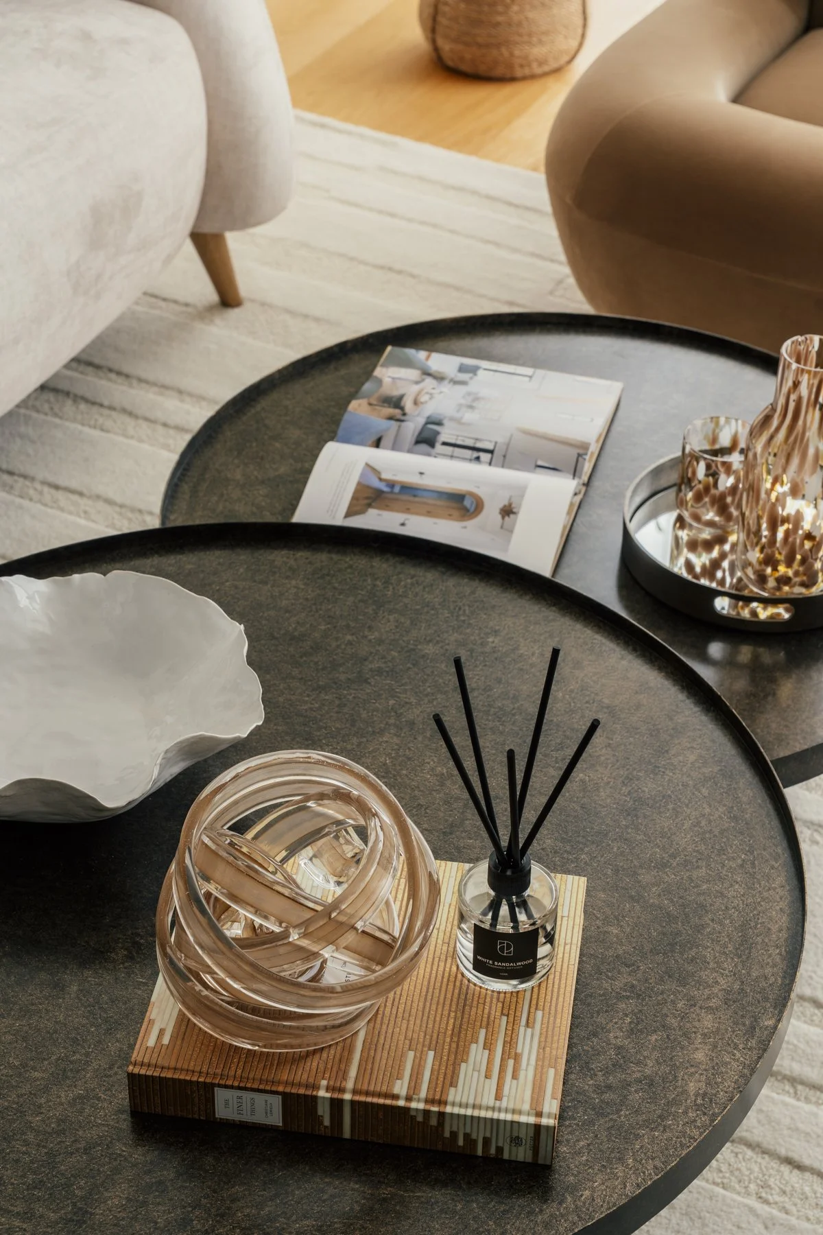 Living room with two black round coffee tables. One table has a book, an open magazine, glass vases, and a reed diffuser. The other table has a white decorative bowl. Beige sofa and armchair, wooden floor, and a beige area rug.