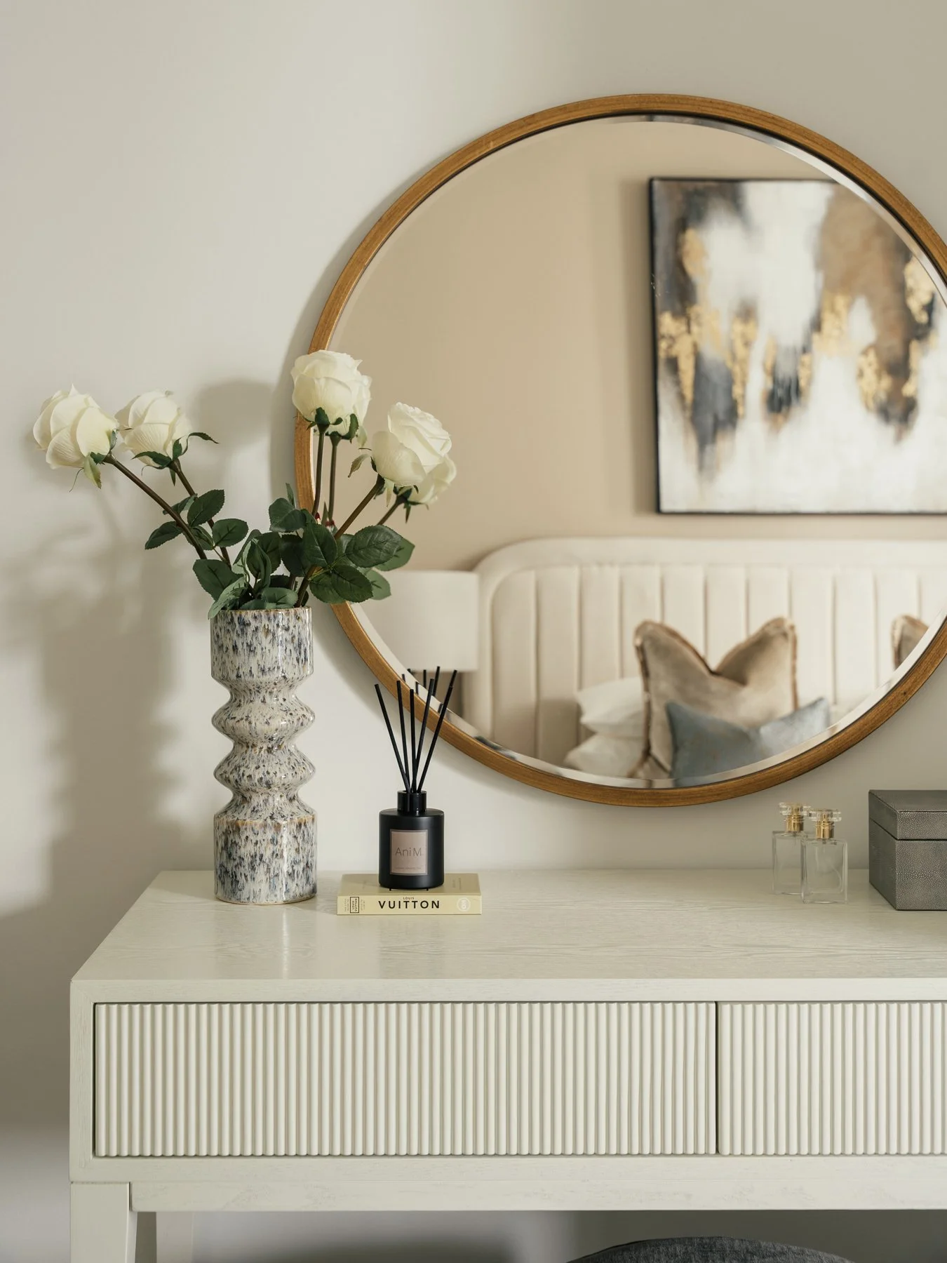 Decorative dresser with a vase of white roses, a reed diffuser, a gray box, and small glass bottles, with a large round mirror and a modern abstract painting on the wall behind.