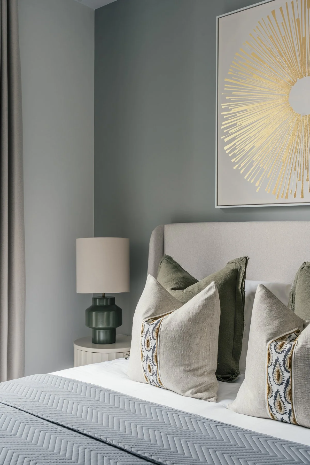 A bedroom with a light grey upholstered bed, green and beige throw pillows, a white bedside table with a dark green lamp, a light grey wall, a large abstract artwork with gold rays, and grey curtains.