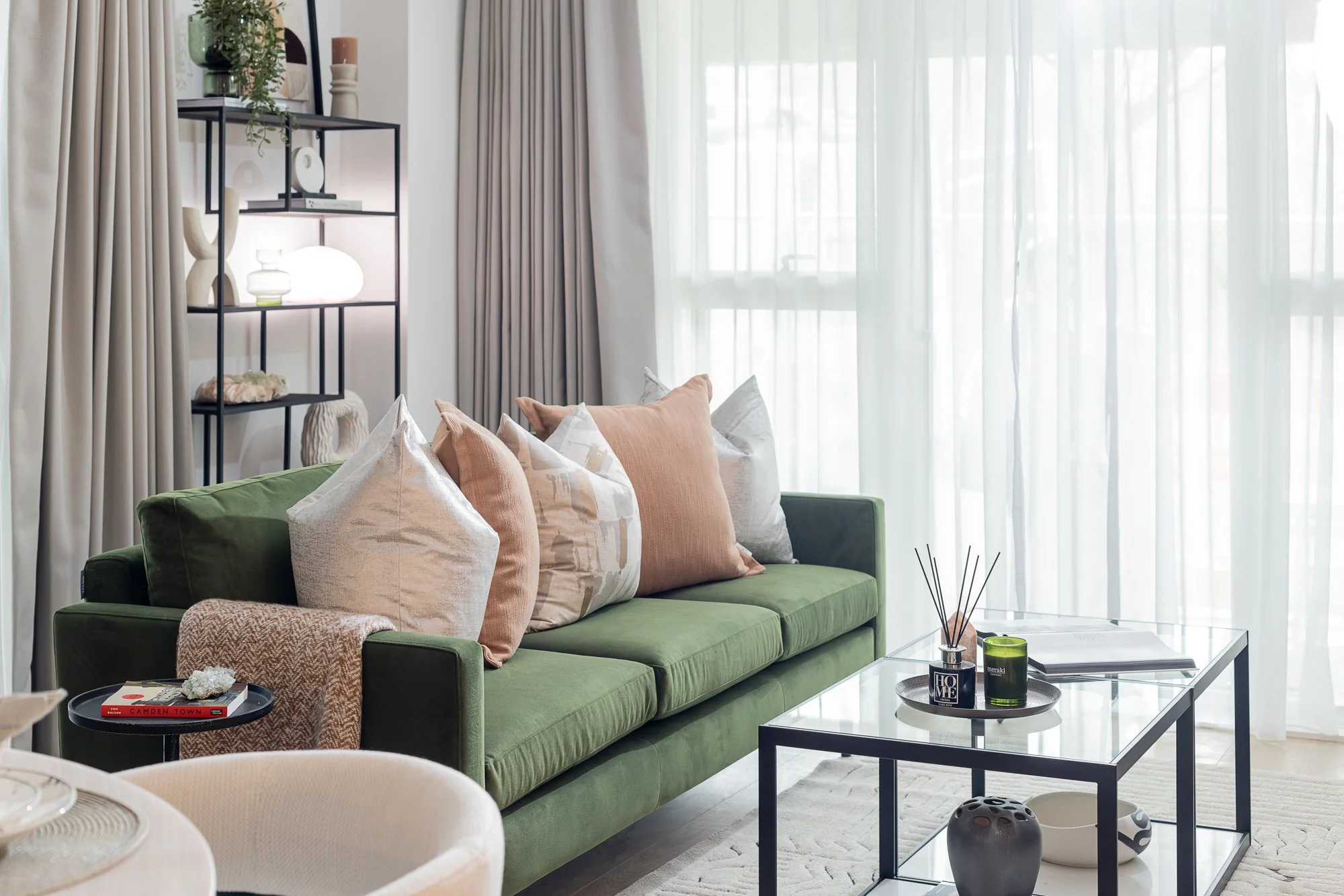 Living room with a green sofa, beige and peach pillows, a glass-top black metal coffee table with decorative items, and a tall black shelving unit with vases, books, and decor, all in front of large windows with sheer white curtains.