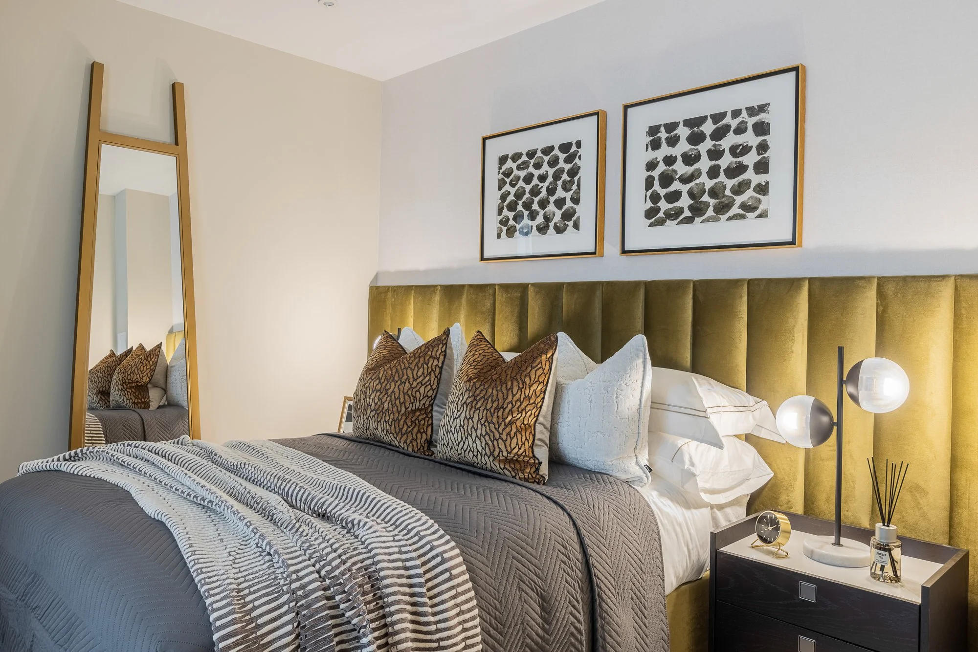 Modern bedroom with a large bed, green velvet headboard, decorative pillows, a nightstand with a lamp, clock, and reed diffuser, framed artwork, and a full-length mirror.