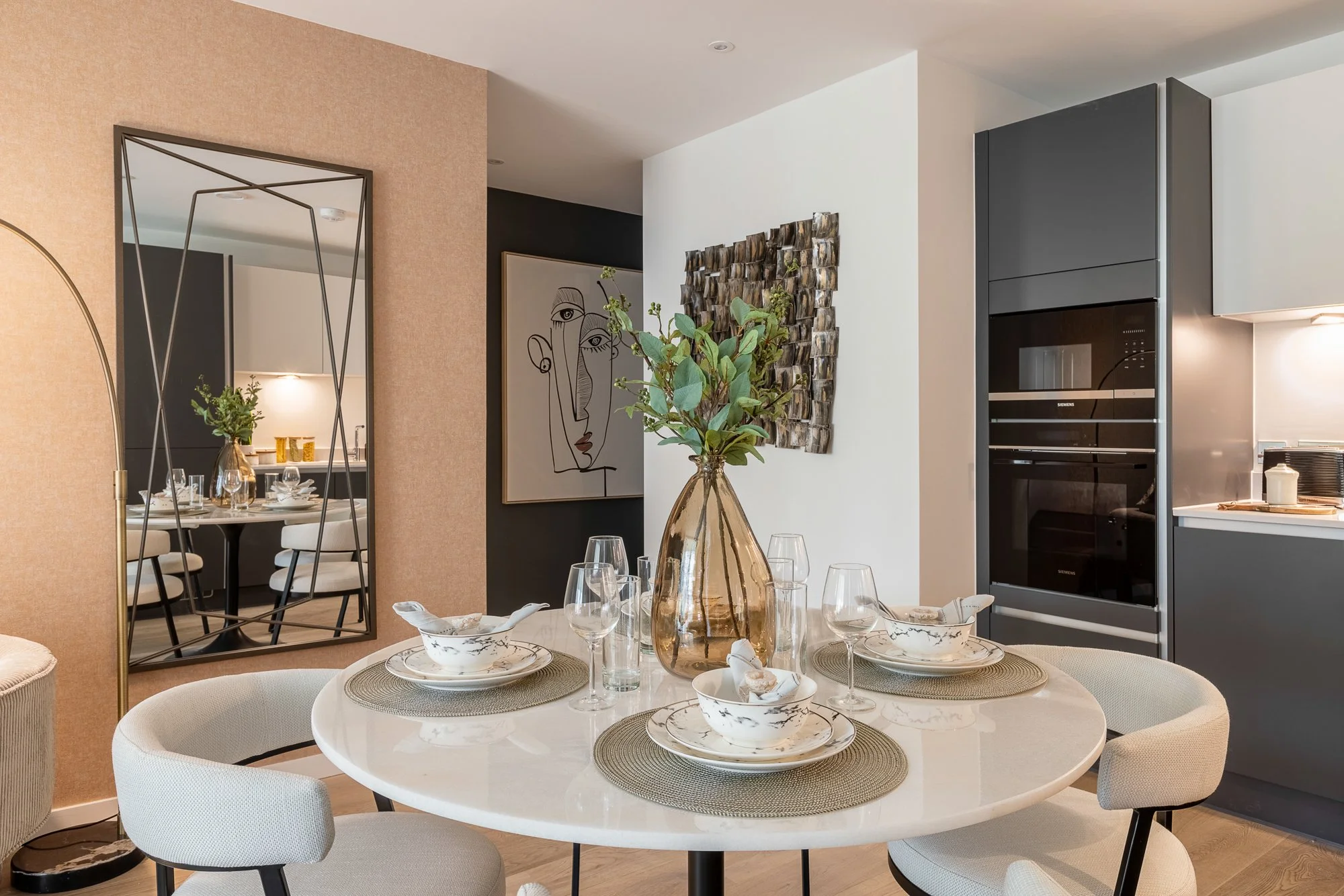 Modern dining area with a round table set with bowls, glasses, and a large vase with green leaves. The background includes a mirror, artwork, a textured wall decor, and a kitchen with black and gray cabinets.