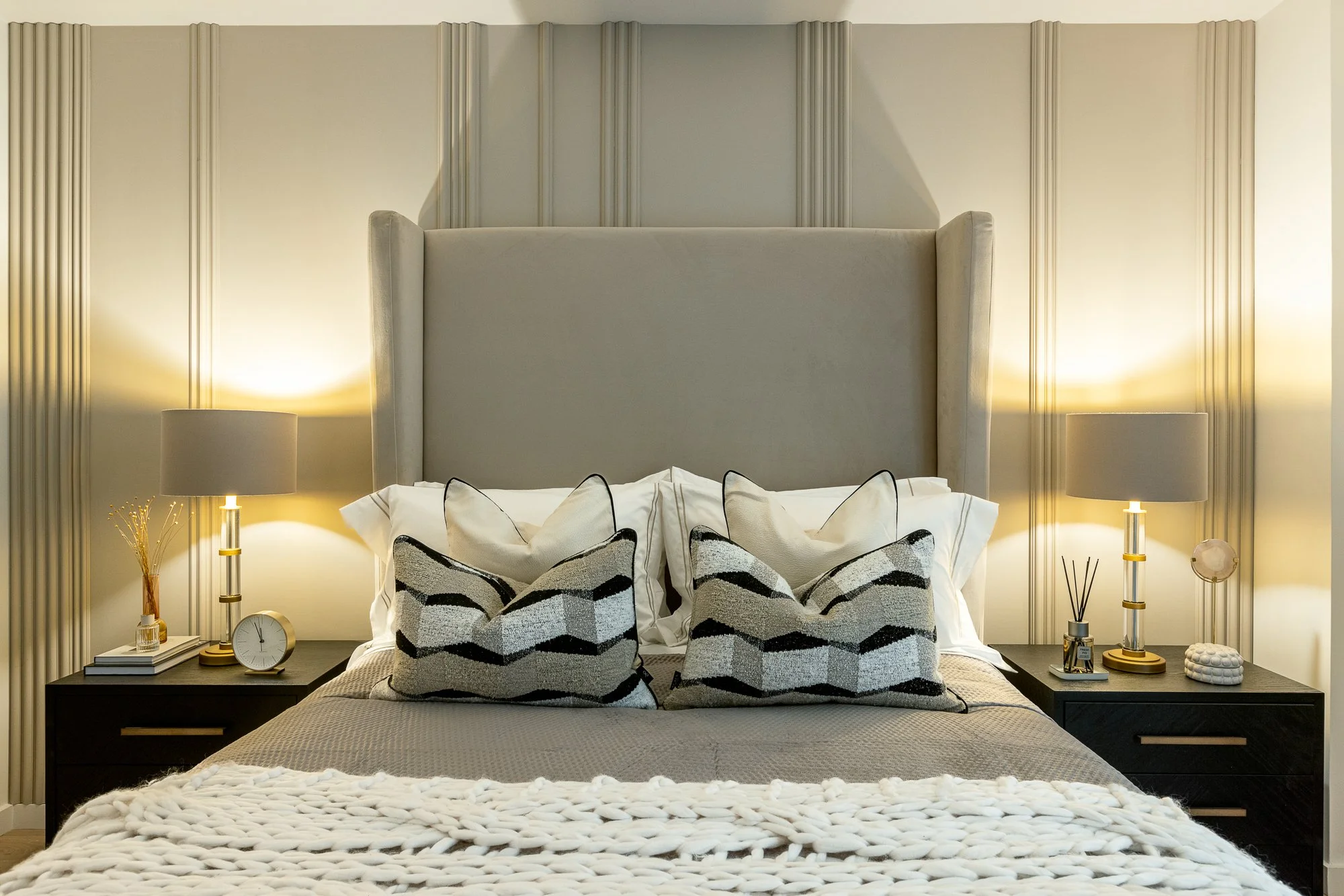 A neatly made bed with beige and black patterned pillows, white sheets, and a chunky beige blanket, flanked by black nightstands with lamps and decorative items, in a modern bedroom with beige paneled wall.