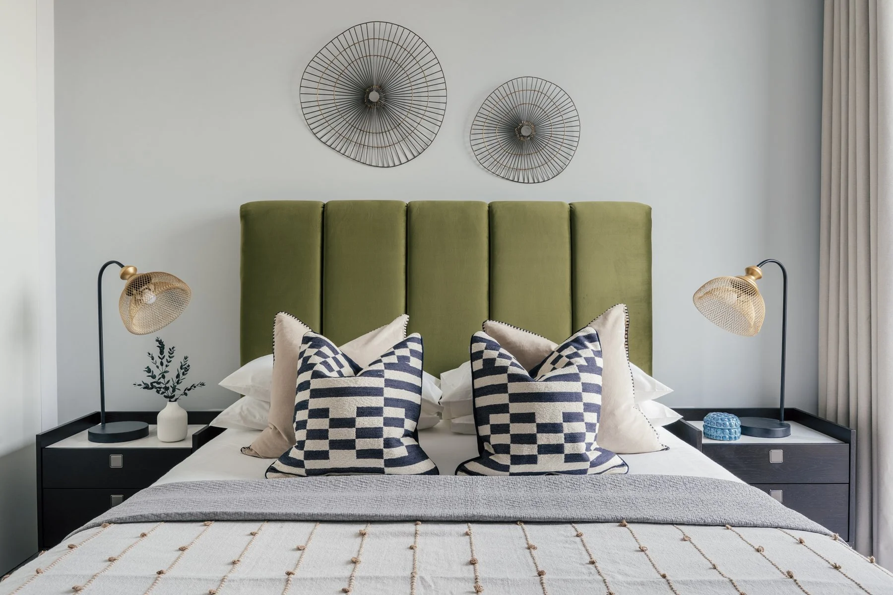 A neatly made bed with a green headboard, four decorative pillows with navy and white checkered patterns, beige pillows, and a blanket. Flanking the bed are black nightstands with modern table lamps, a small plant in a white vase, and a blue decorati