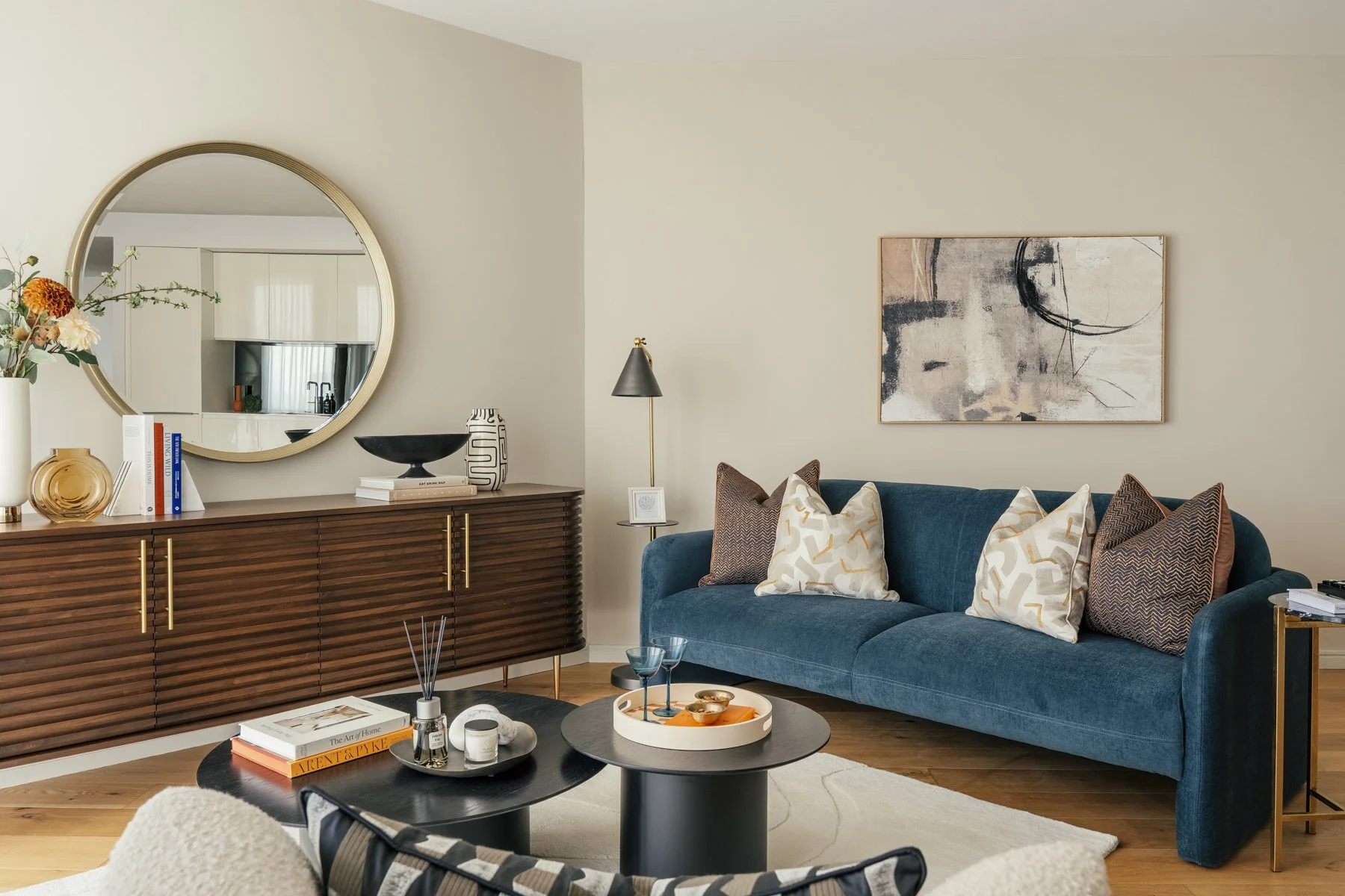 Living room with a blue sofa, patterned and solid throw pillows, a side table, a wall art, a black coffee table with tray and drinks, brown wood sideboard with decor and mirror, and hardwood floors.