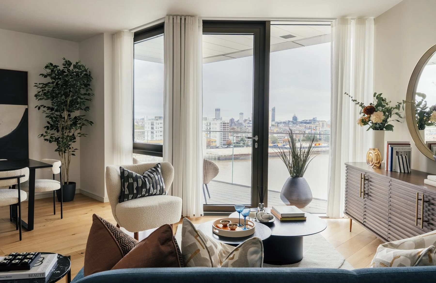 Modern living room with large glass doors leading to a balcony, city skyline view, light-colored wood flooring, white curtains, sofa with cushions, round coffee table with decorative items, armchair with striped cushion, tall potted plant, sideboard 