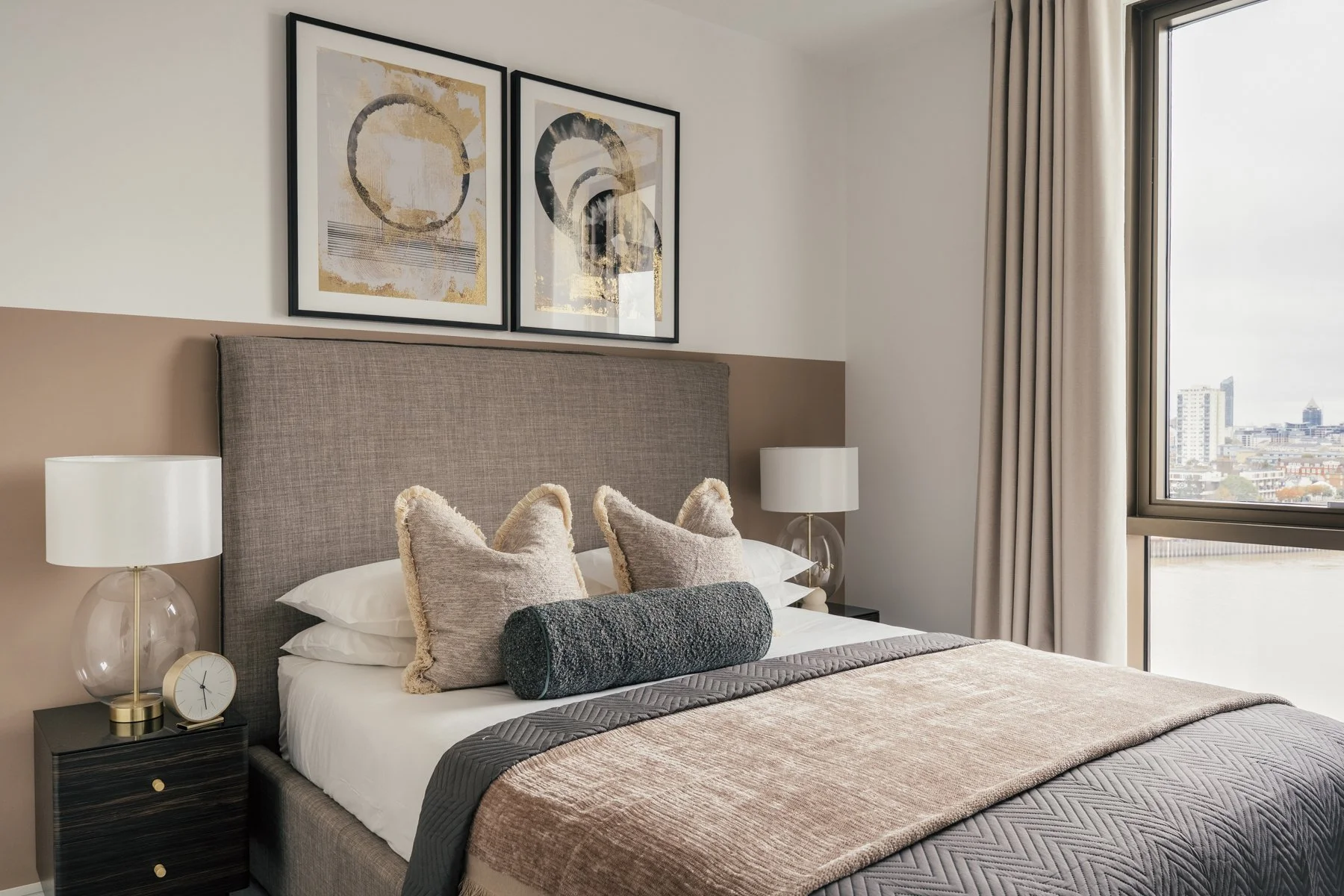 Modern bedroom with a large bed, beige headboard, two table lamps, and decorative pillows, with city skyline view through a large window.
