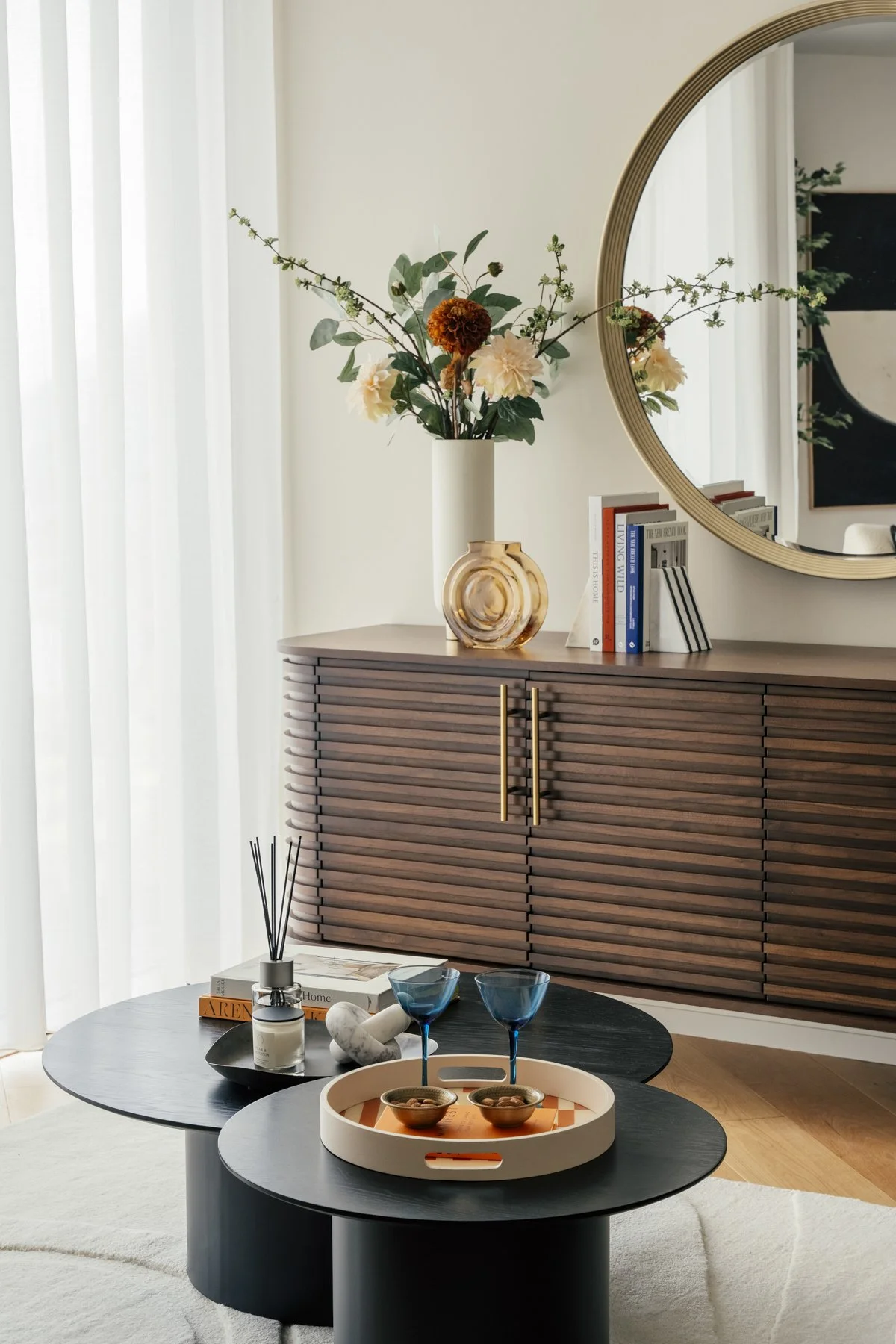 Living room with a black round coffee table with two blue wine glasses and a tray, a wooden sideboard with a large round mirror, a white vase with flowers, and books. Draped curtains and part of a white rug on a wooden floor are also visible.