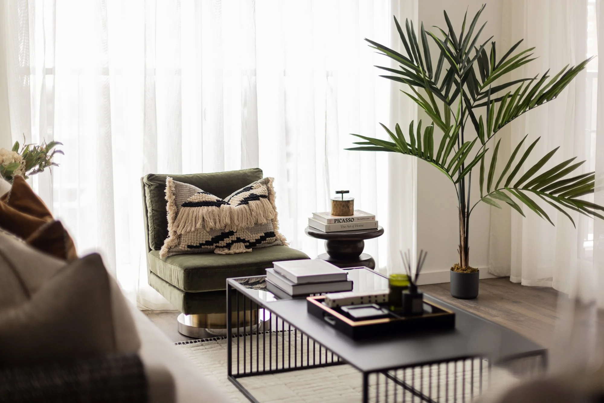 A modern living room with sheer white curtains, a dark green armchair with a patterned cushion, a black coffee table with books and decor, a side table with a stack of books, and a large potted palm plant.