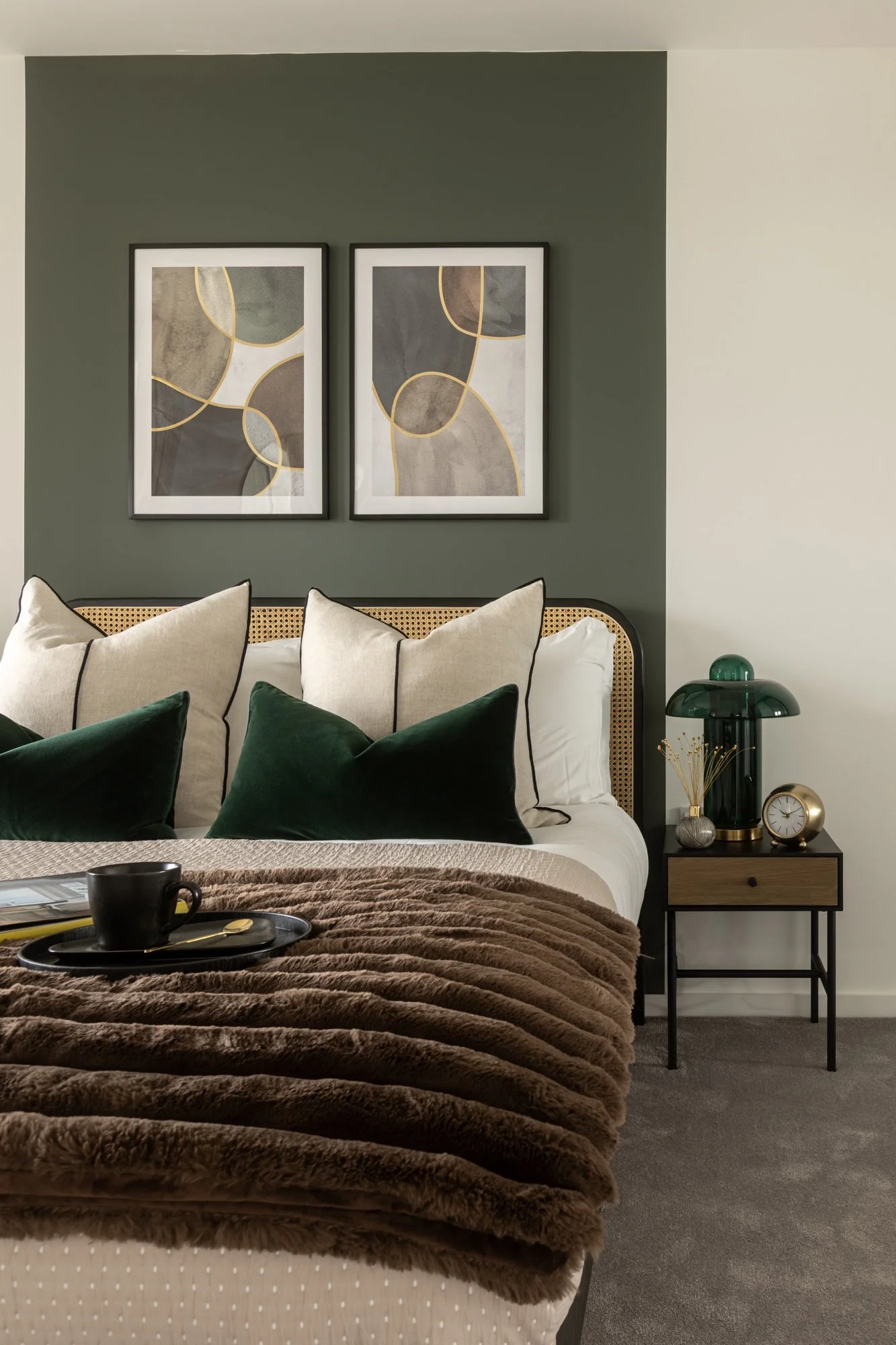 A cozy bedroom with a bed featuring beige and green pillows, a plush brown blanket, and modern decor including wall art, a green glass lamp, and a small bedside table with a clock and decorative items.