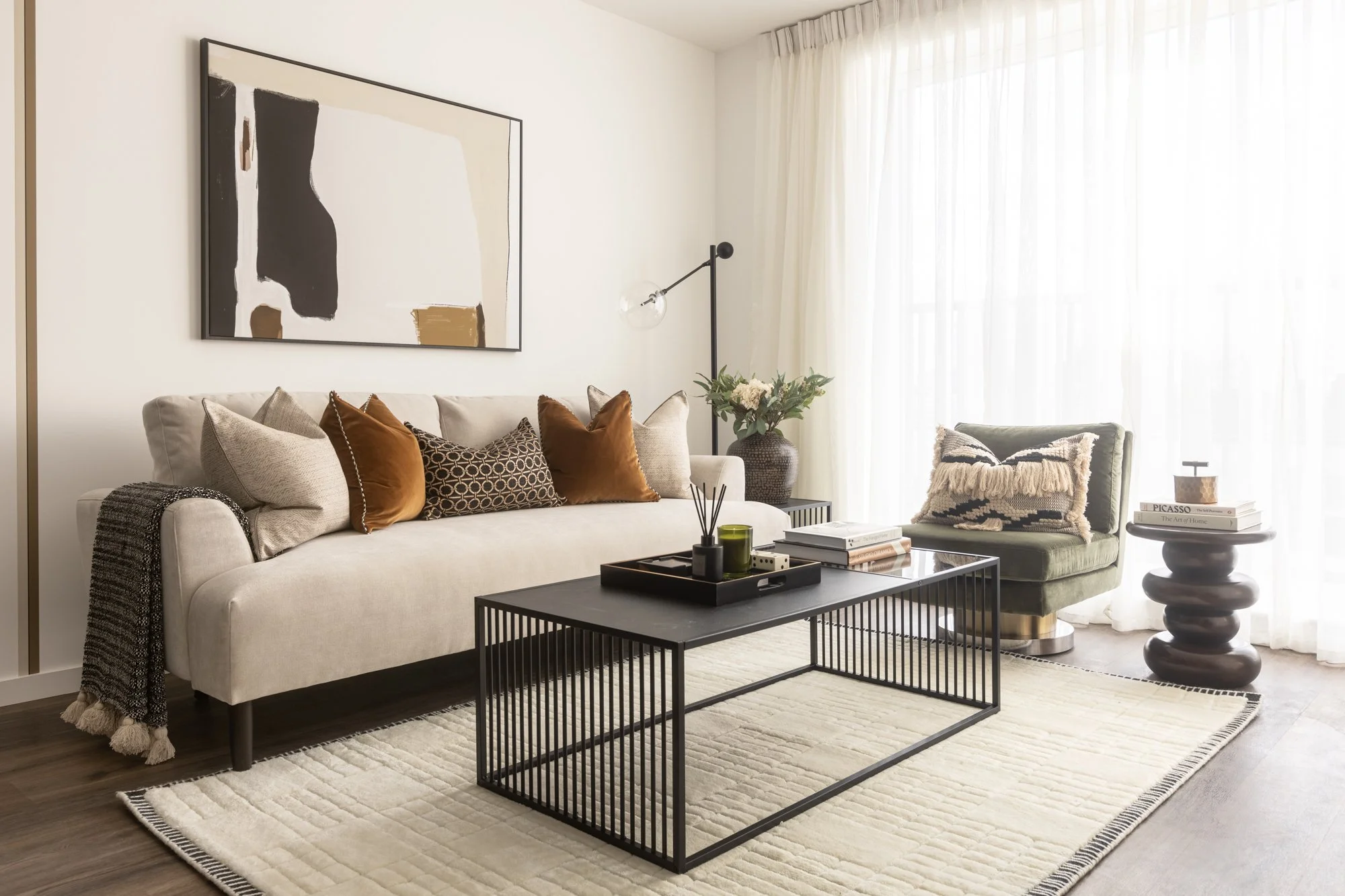 Living room with a beige sofa and colorful pillows, a black coffee table with decorative items, a green armchair with a textured pillow, a side table with books, large window with white curtains, and abstract wall art.