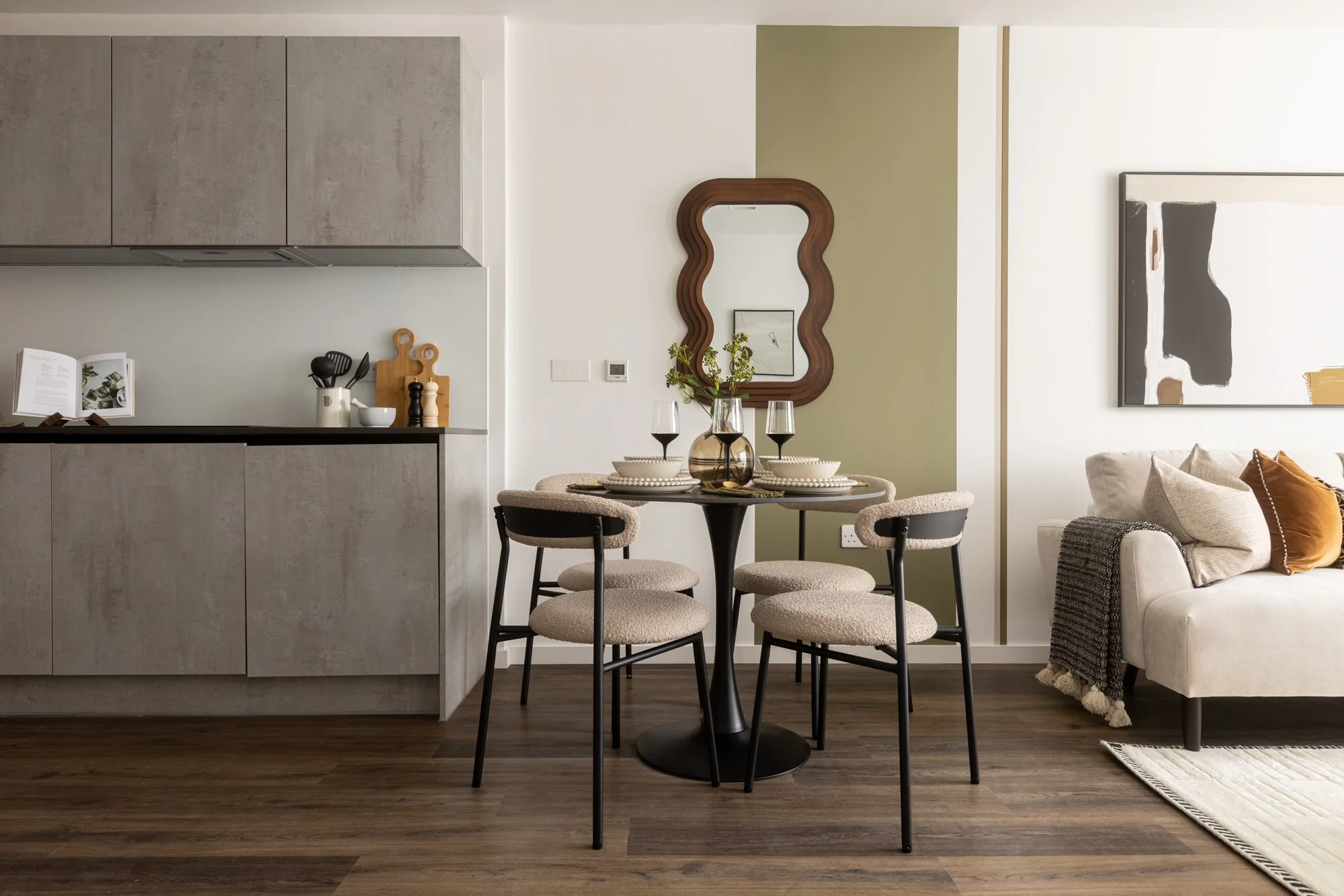 Modern living and dining area with a round table, four beige upholstered chairs, a gray kitchen cabinet, a decorative mirror, and a white sofa with pillows