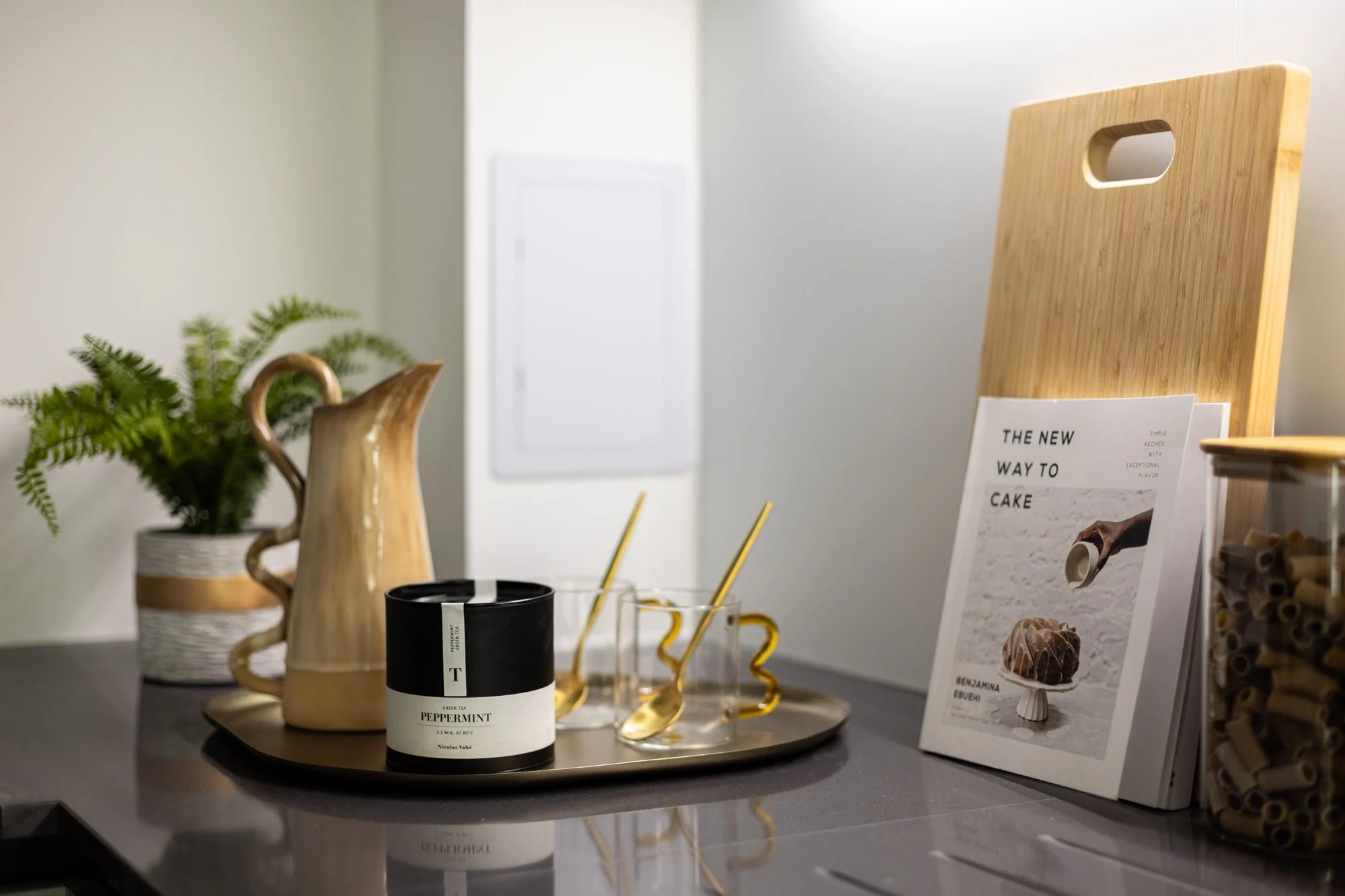 Decorative kitchen counter with a fern plant in a pot, a wooden pitcher, a tray holding a Peppermint green tea candle, two gold-colored spoons and two clear cups with gold-colored straws, a wooden cutting board holding a book titled "The New Way to C