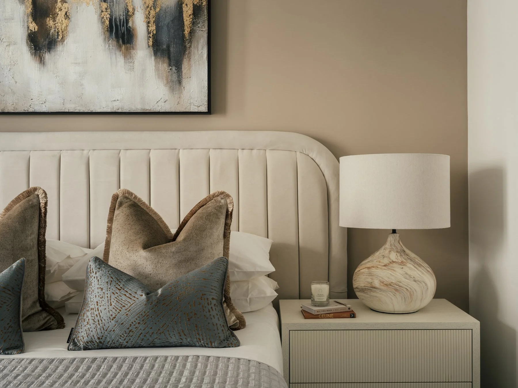 Close-up of a beige bed with decorative pillows in front of a white headboard, a beige wall with an abstract painting above, and a cream-colored bedside table with a large marbled ceramic lamp, a candle, and a few books.