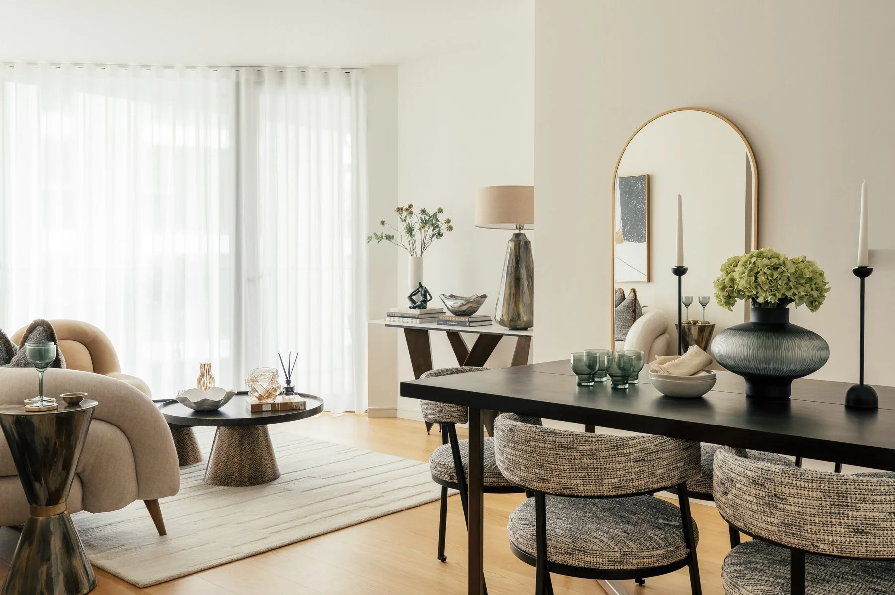 Modern living room with beige sofa, black coffee table, and large windows with sheer white curtains. Dining area with a black table, patterned chairs, decorative vases, and candles.