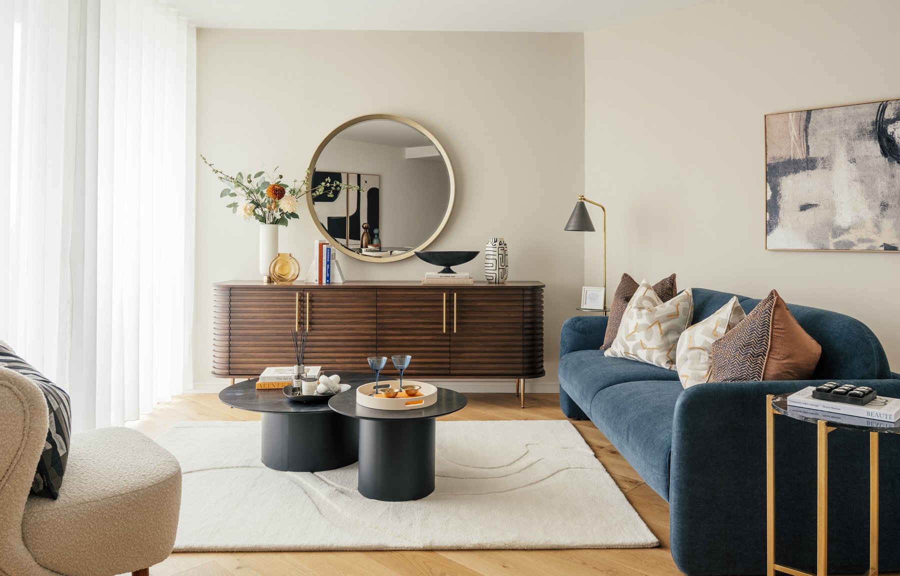 Modern living room with a blue velvet sofa, a white area rug, and dark coffee tables with decorative items. A wooden sideboard with a round mirror, vases, books, and a black bowl is against the wall. There's a lamp with a black shade and gold stand n