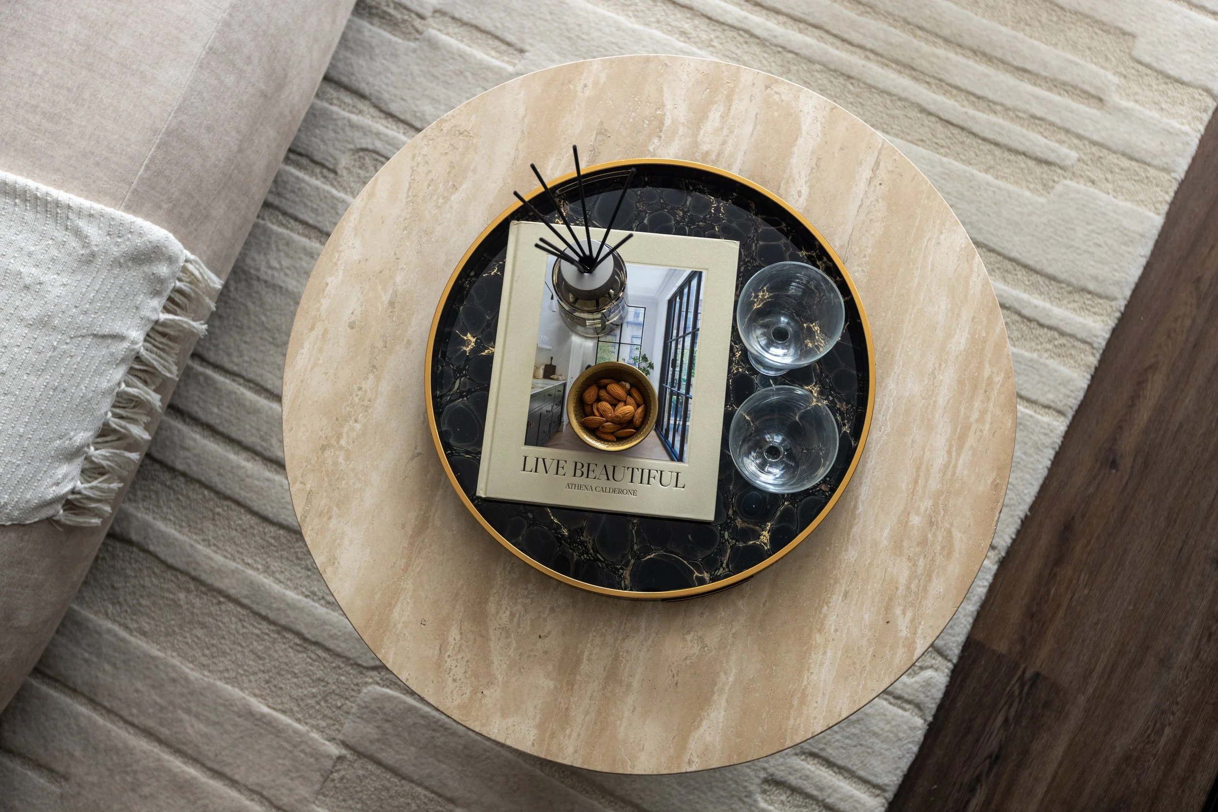 Round beige marble side table with a black and gold tray on top. The tray holds two empty glasses, a small bowl of almonds, a book titled "Live Beautiful" by Athena Calderone, and a reed diffuser. The table is next to a light-colored sofa with a textured pillow, and the floor is brick with a rug and a wooden floor nearby.