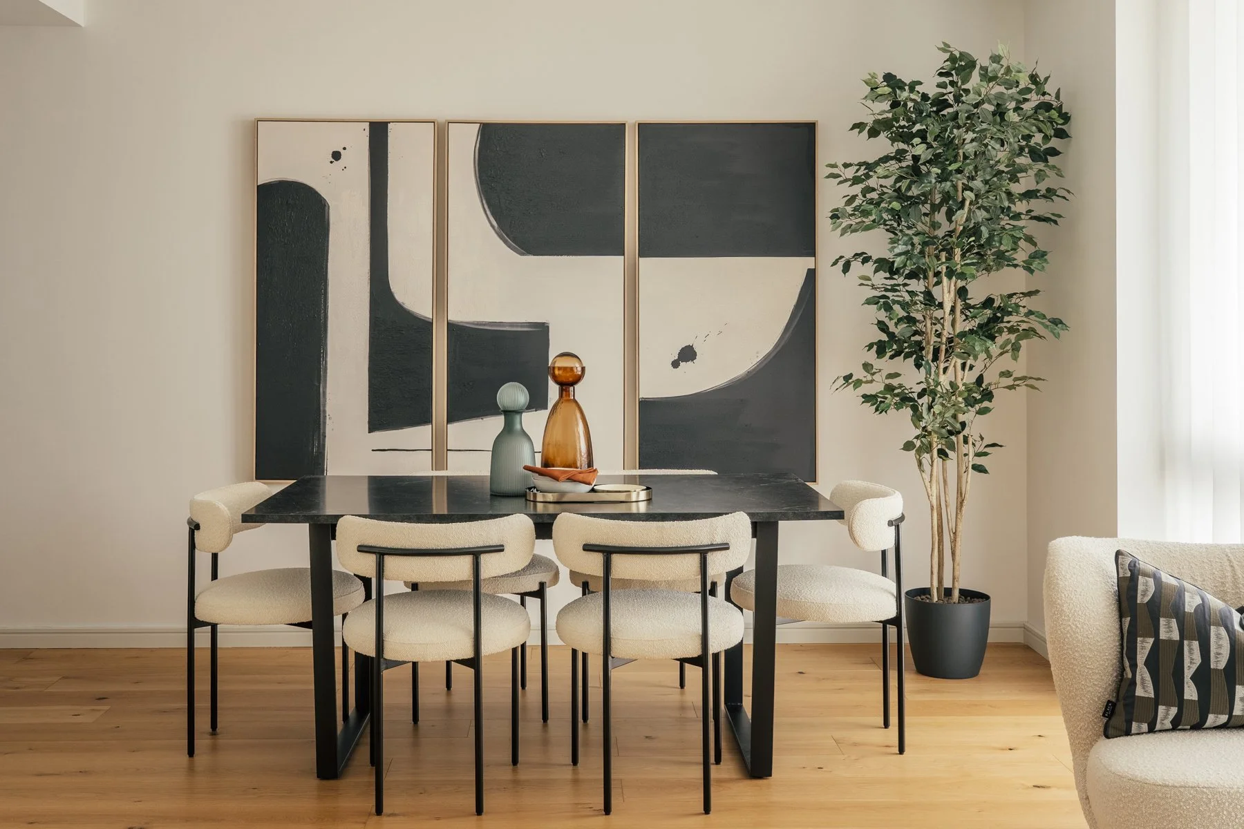 Modern dining room with black table, six cream-colored chairs, a large abstract black and white painting on the wall, a potted tree, and a beige armchair with a decorative pillow, hardwood floors, and natural light.