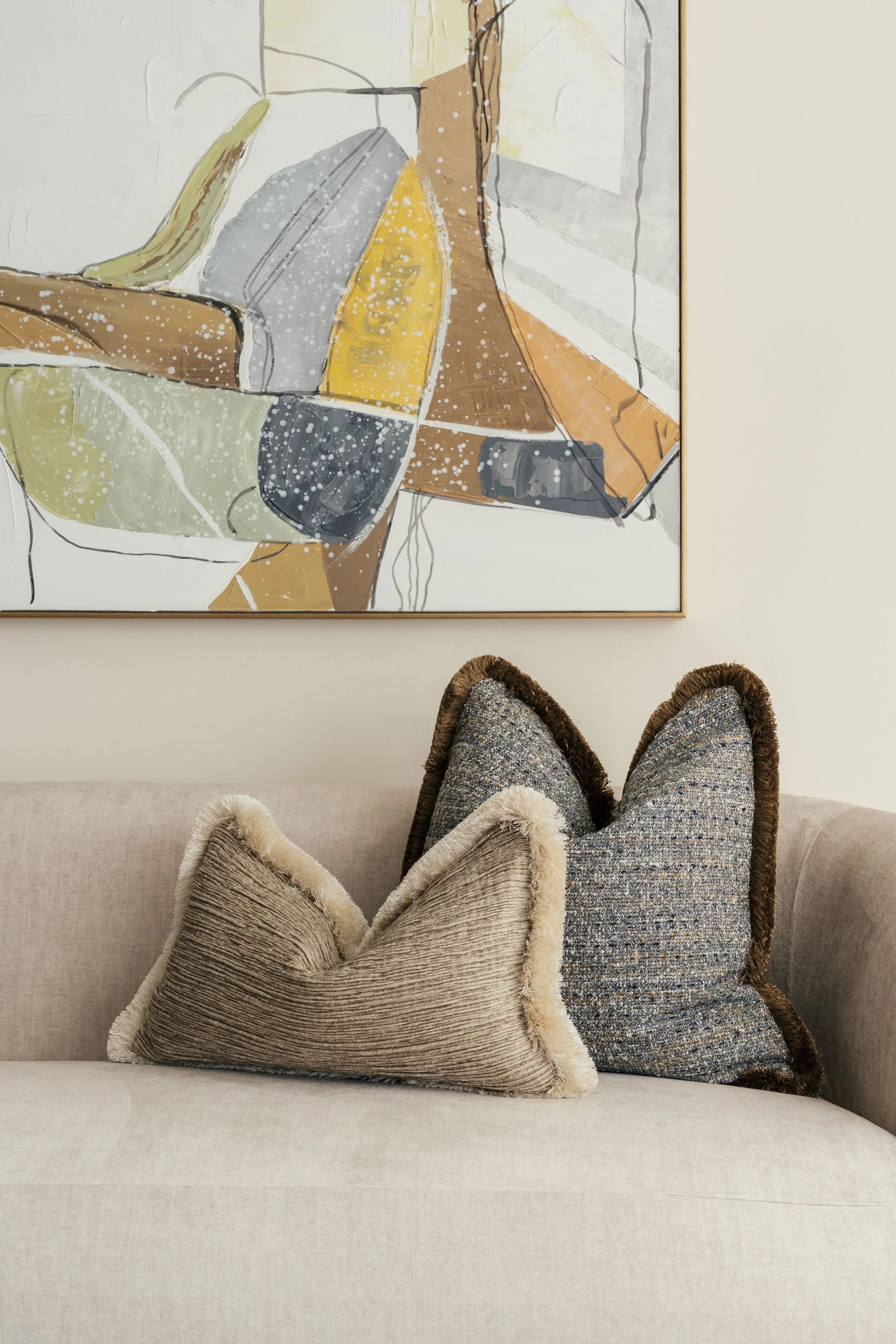 A beige sofa with two decorative pillows, one with a furry edge, topped with a larger dark gray pillow, in front of a beige wall and a large abstract painting with various colors.