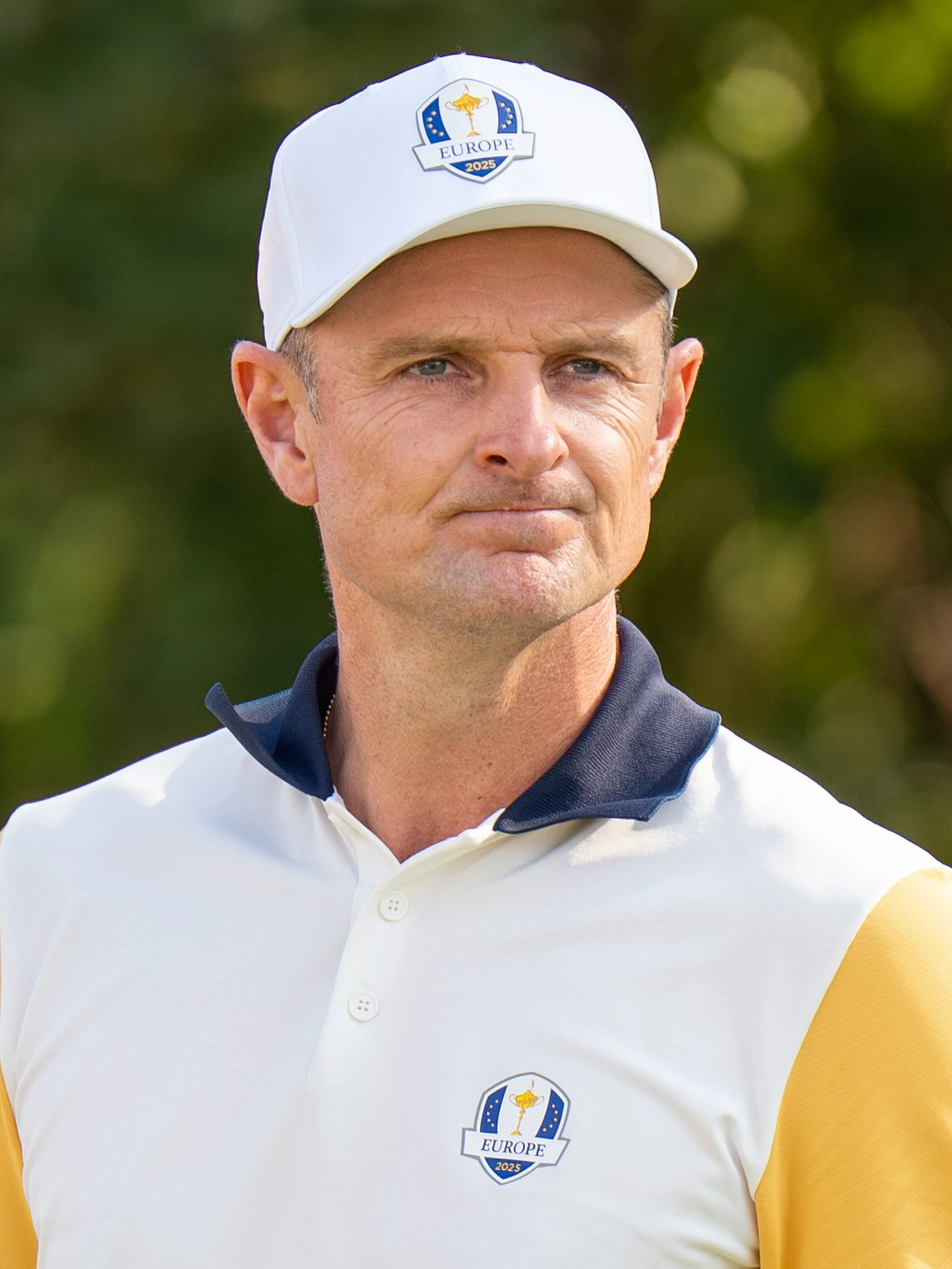 A man wearing a white golf cap with a logo that says 'Europe 2025' and a white golf shirt with yellow and black accents, standing outdoors with blurred greenery in the background.