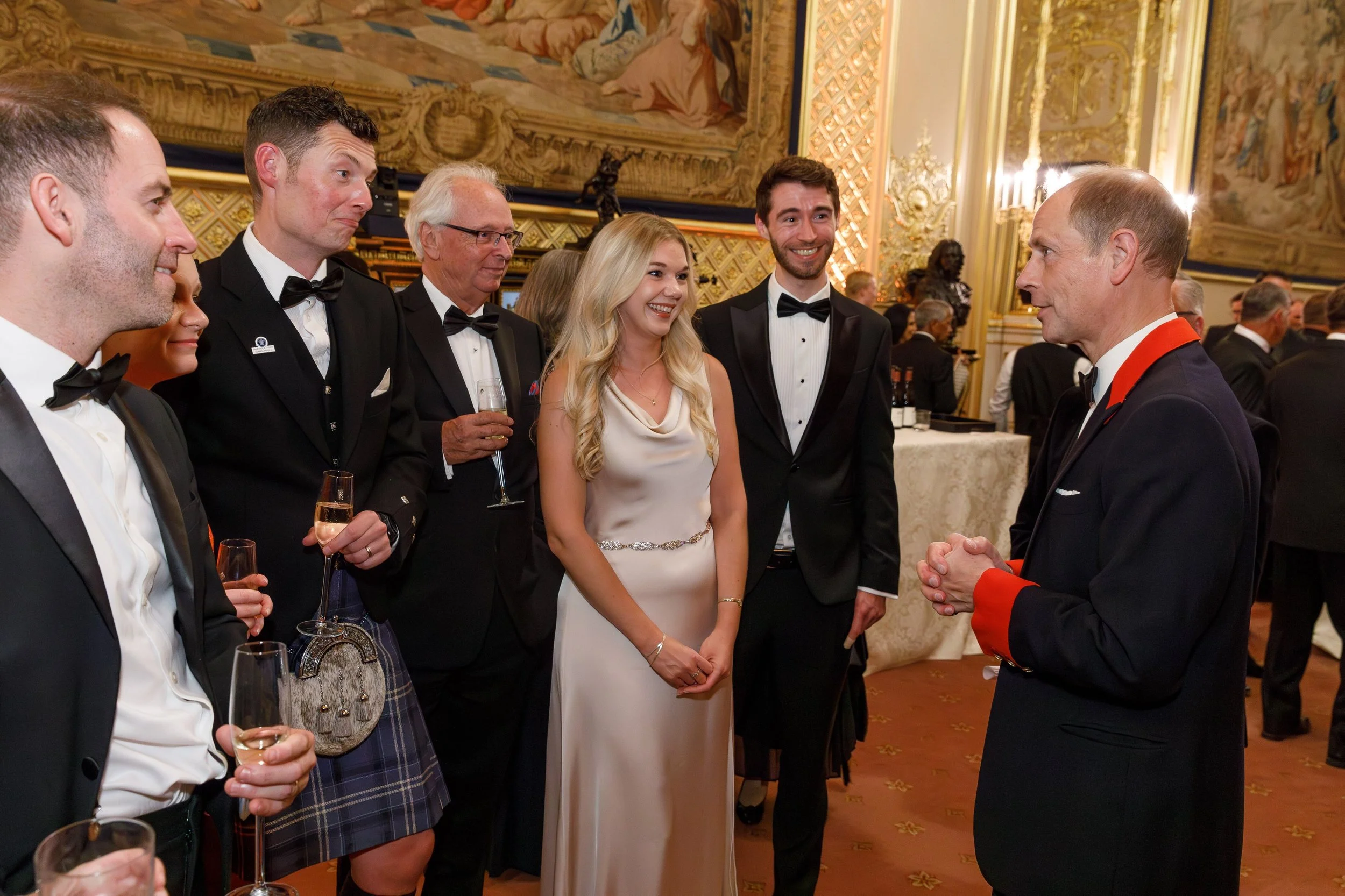 A group of well-dressed people at a formal event, including men in tuxedos and a woman in a satin gown, engaging in conversation with a man in a dark suit with red accents, inside a decorated, elegant hall.