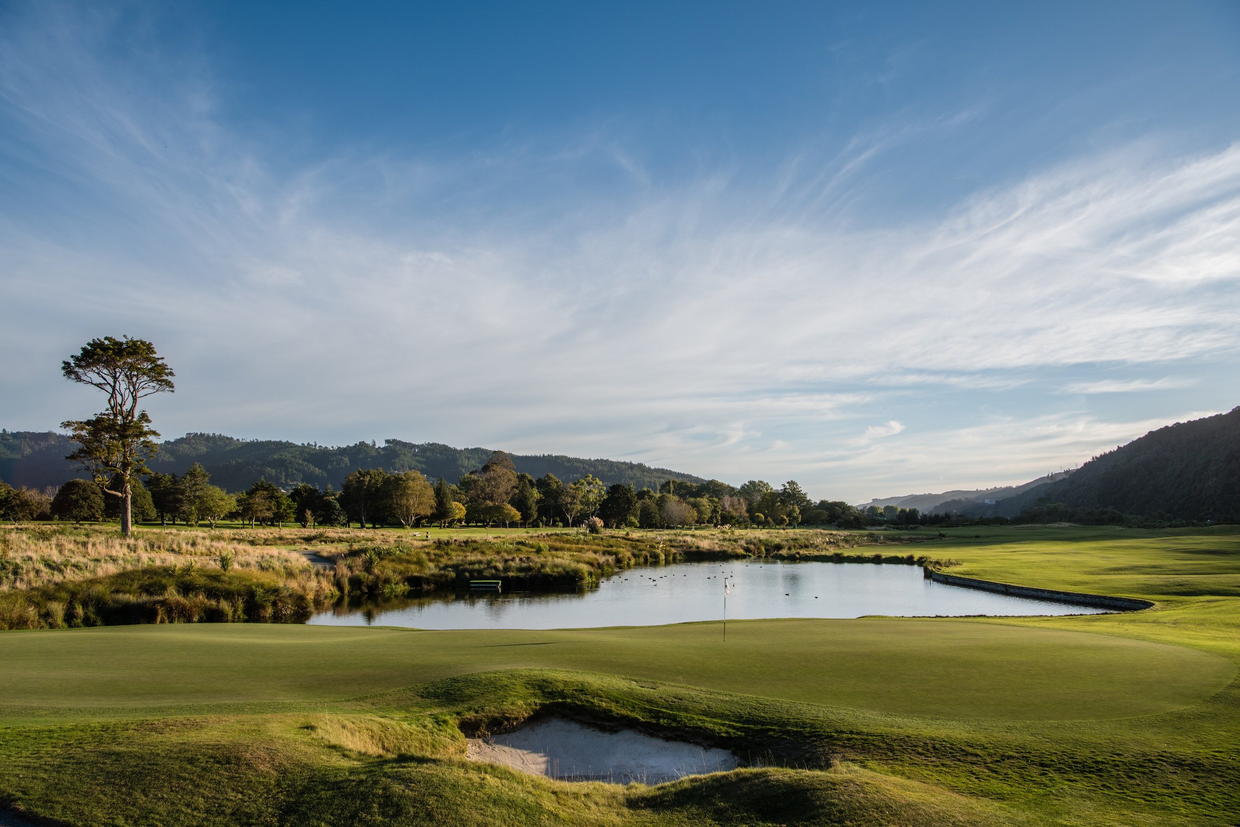 A scenic golf course with lush green grass, a small pond, and a backdrop of trees and rolling hills under a partly cloudy sky.