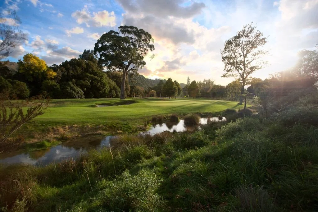 A scenic golf course with lush green grass, surrounded by trees and bushes, with a small pond reflecting the sky, under a partly cloudy sky with the sun setting or rising.